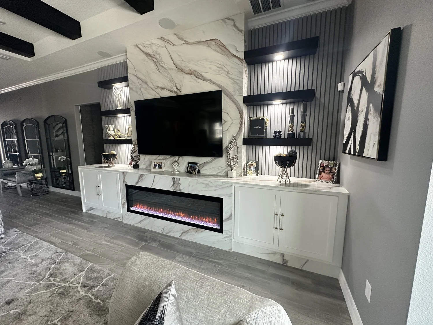 Modern living room with a marble wall feature, flat-screen TV, electric fireplace, gray flooring, and decorative items on white cabinets and black shelves.