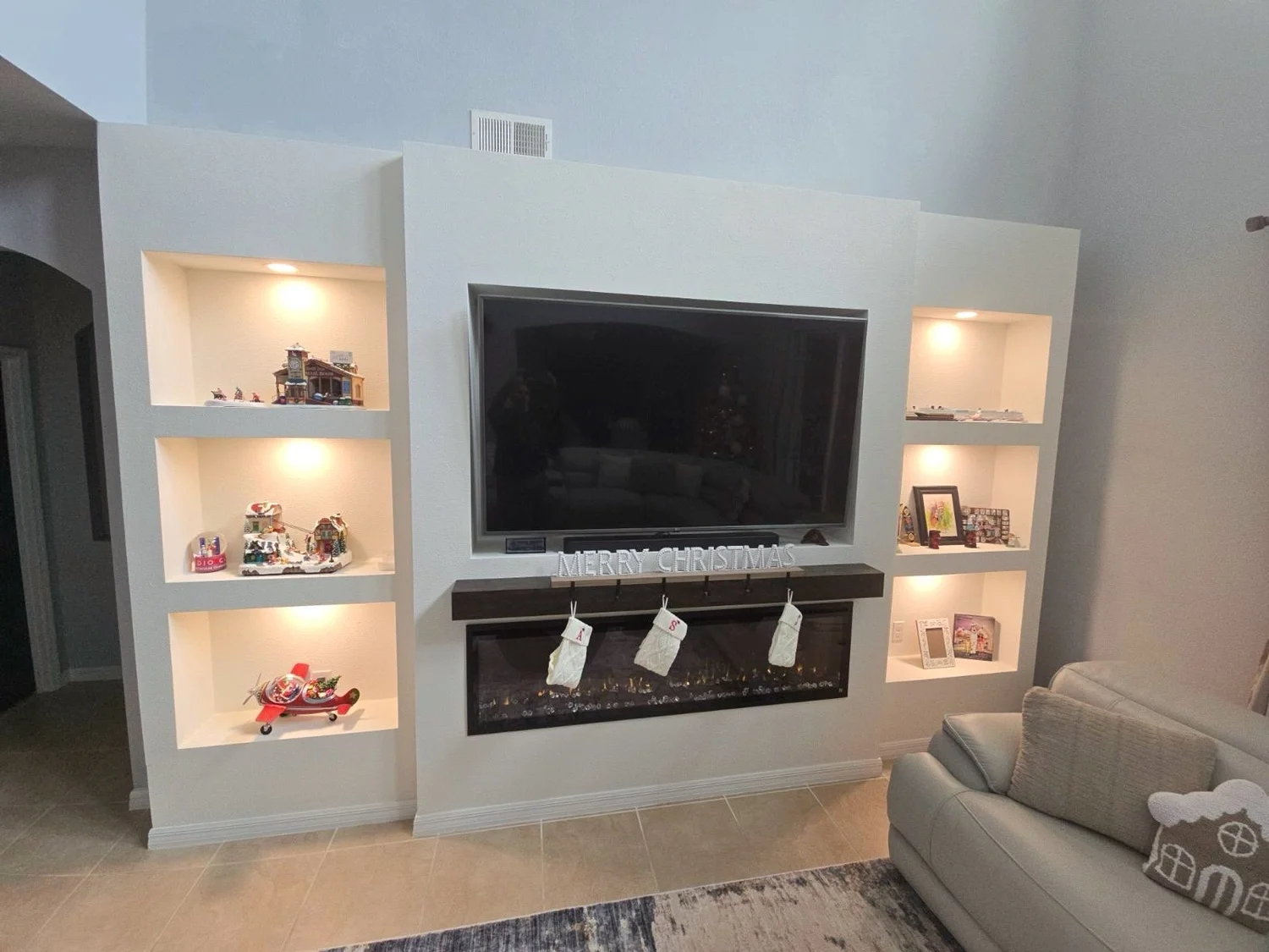 Living room wall with built-in shelves displaying Christmas decorations, a flat-screen TV, and a modern fireplace with a 