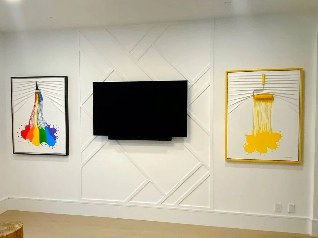 Minimalist living room wall with a black flat-screen TV mounted in the center, two colorful paint splatter artworks on each side, one with rainbow colors and the other yellow, and a white wall with geometric paneling.