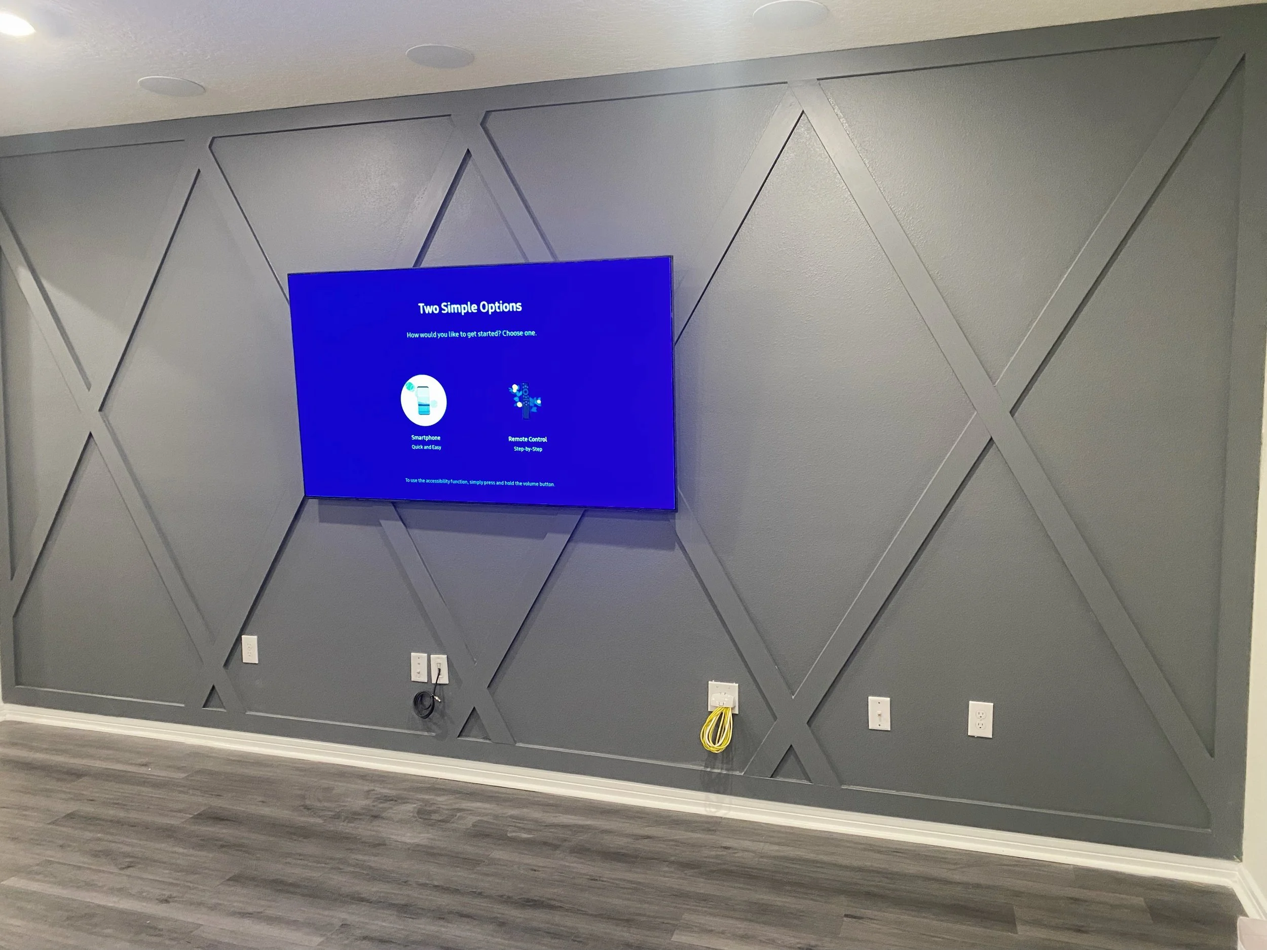 A wall with gray textured panels and diagonal lines, a mounted flat-screen TV displaying a blue screen with options, electrical outlets, and yellow cables on the lower part of the wall.