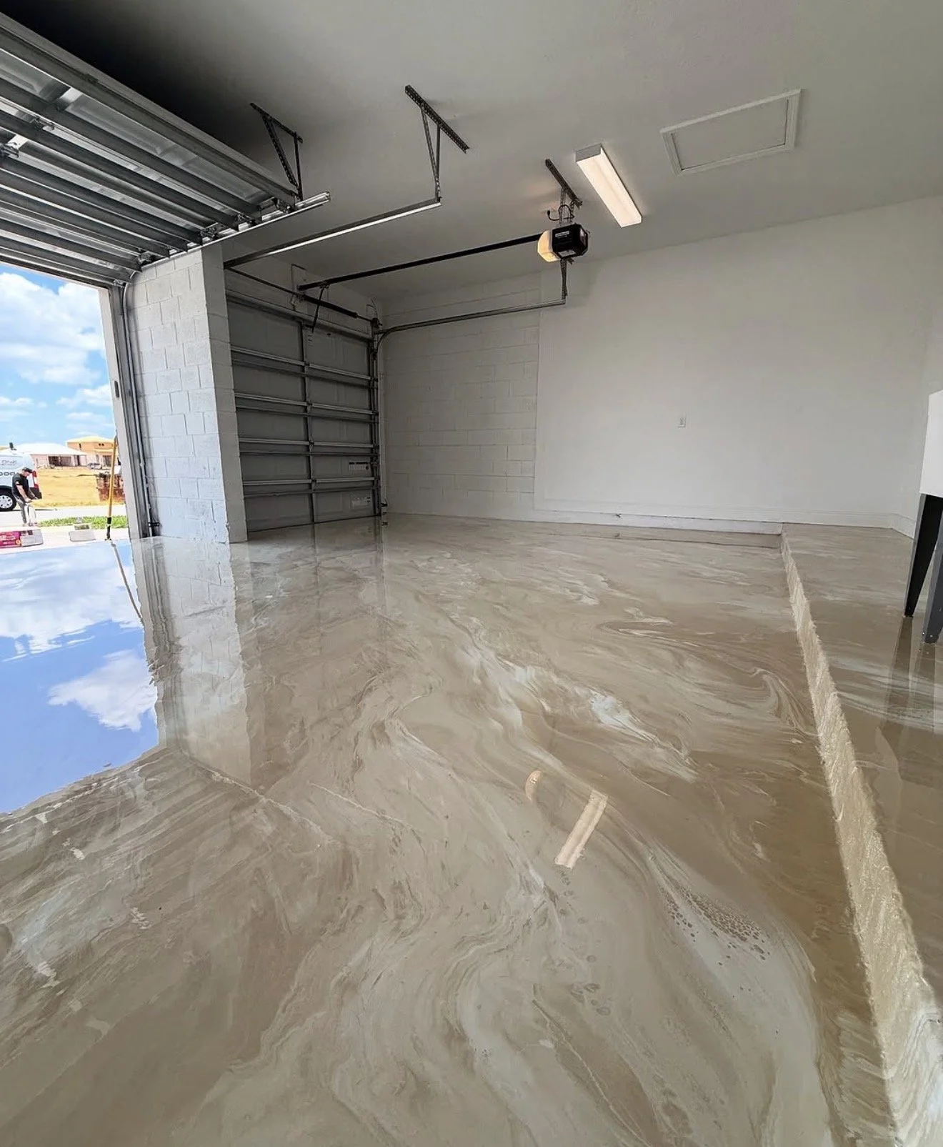 Empty garage with polished marble floor, partially open garage door, and white walls. Outside, a person is visible near a vehicle.