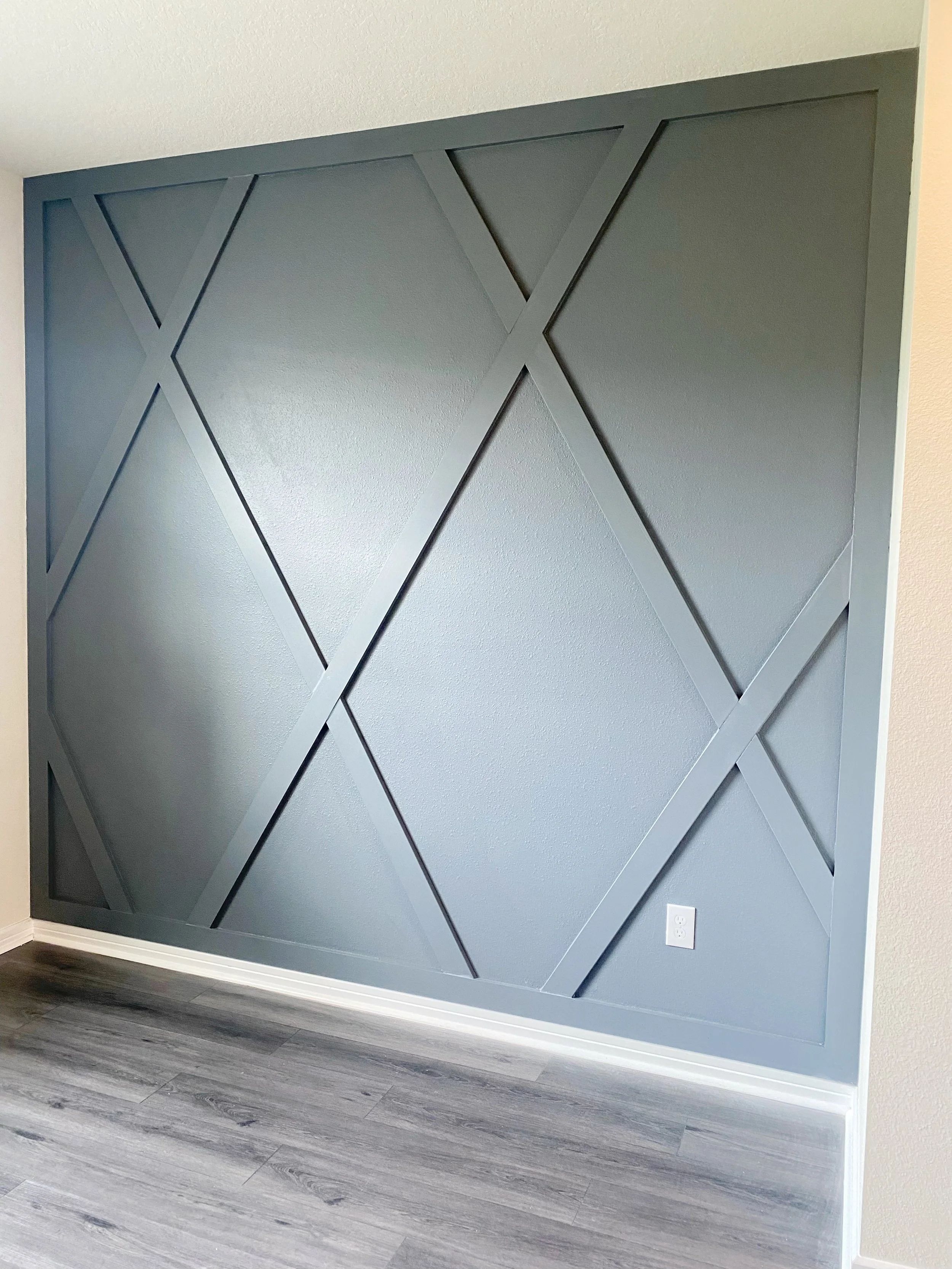 Interior wall with decorative gray panel featuring geometric patterns, located near a wood-like flooring and an electrical outlet.