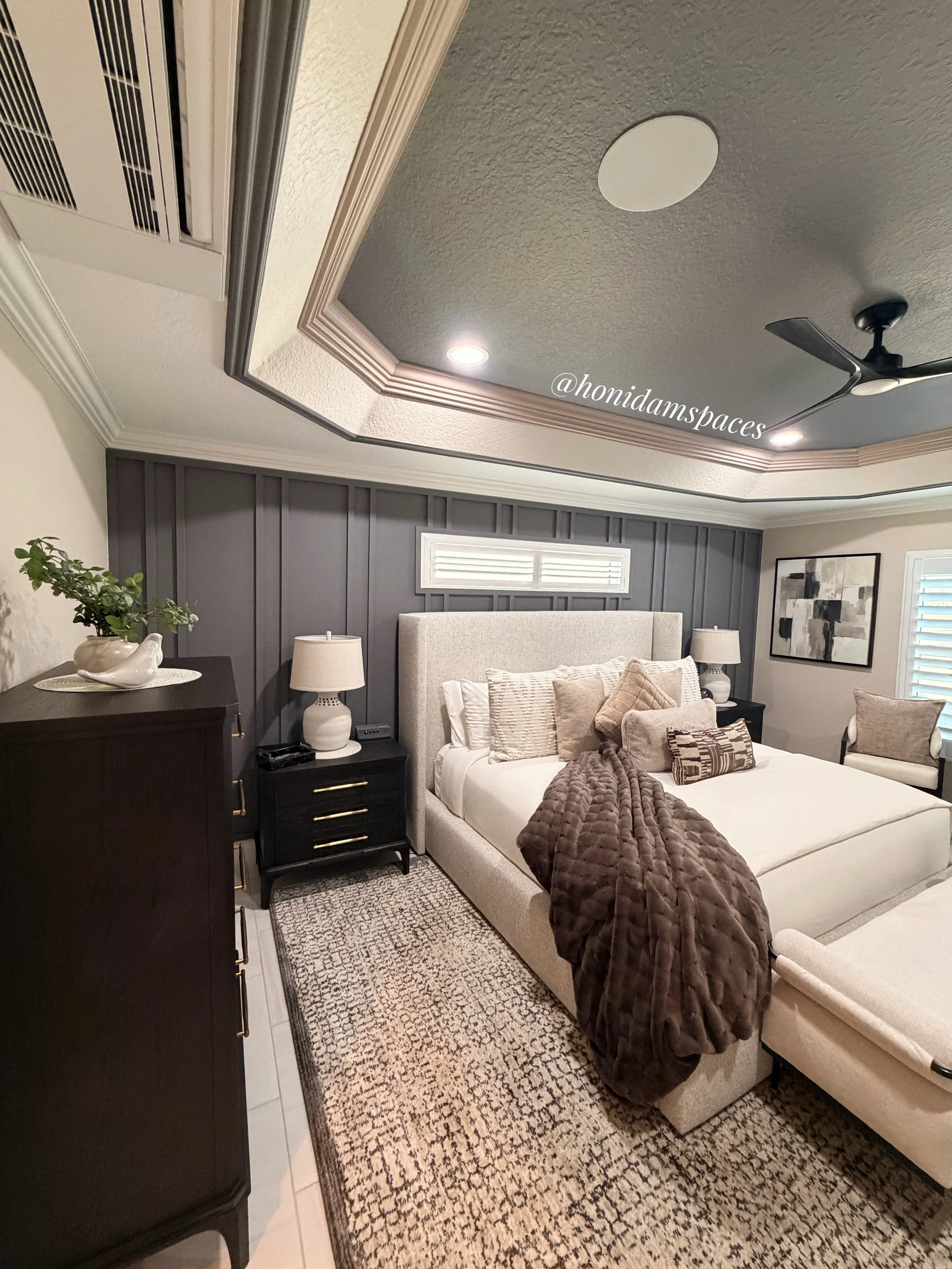 A bedroom with a bed, dark wood nightstand, lamps, artwork, dark gray paneled wall, window with shutters, ceiling fan, and decorative rug.