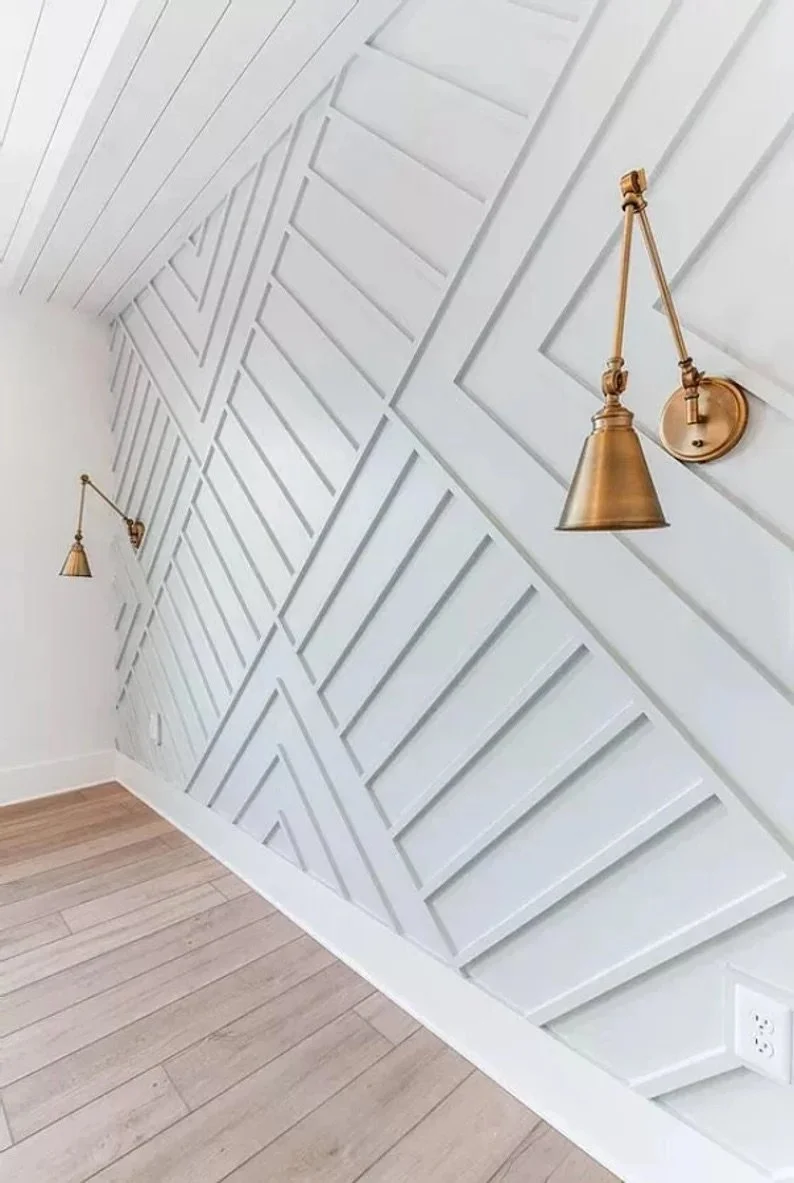 Interior corner of a room with a diagonally designed white wooden wall paneling and two gold wall sconces with adjustable arms, beige walls, and light wood flooring.