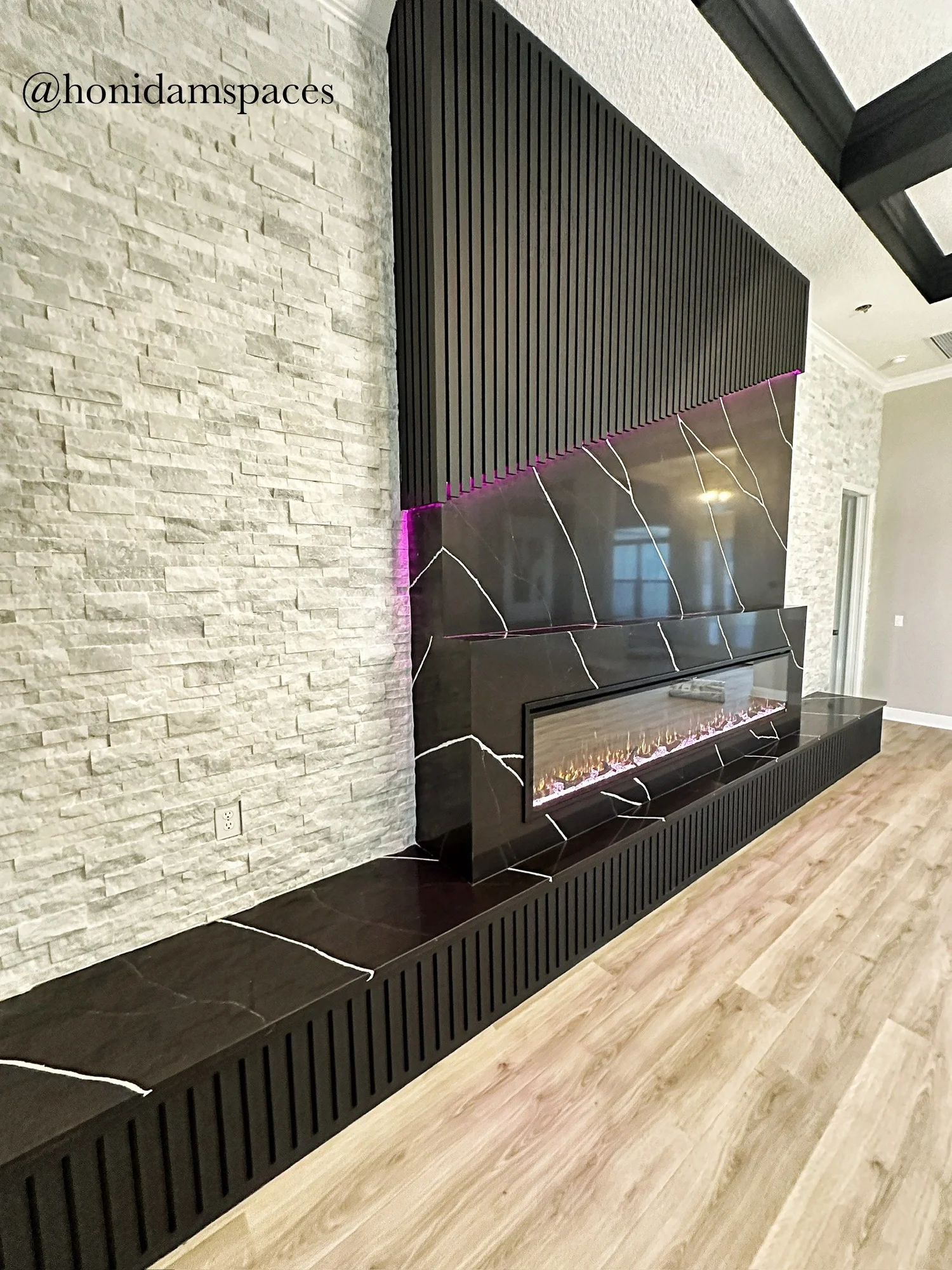 Modern living room fireplace with black marble and white veining, set against a textured white stone wall, with a black wooden slatted panel above and a matching vent base, hardwood flooring.