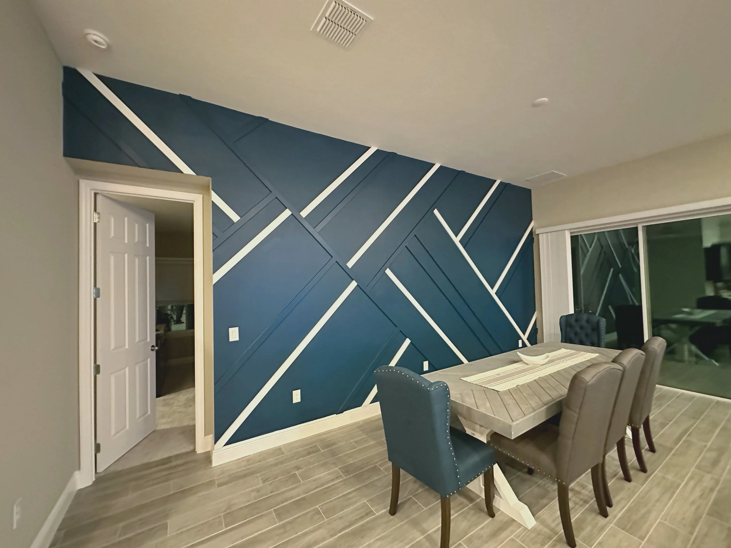 Dining room with a blue geometric accent wall, a wooden table, and six upholstered chairs. Sliding glass door reveals an outdoor patio area.