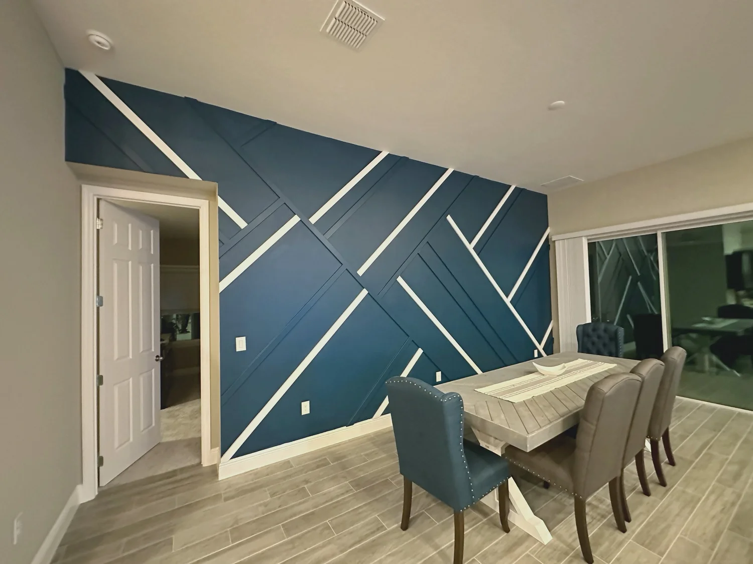 Dining room with a blue geometric accent wall, a wooden table, and six upholstered chairs. Sliding glass door reveals an outdoor patio area.