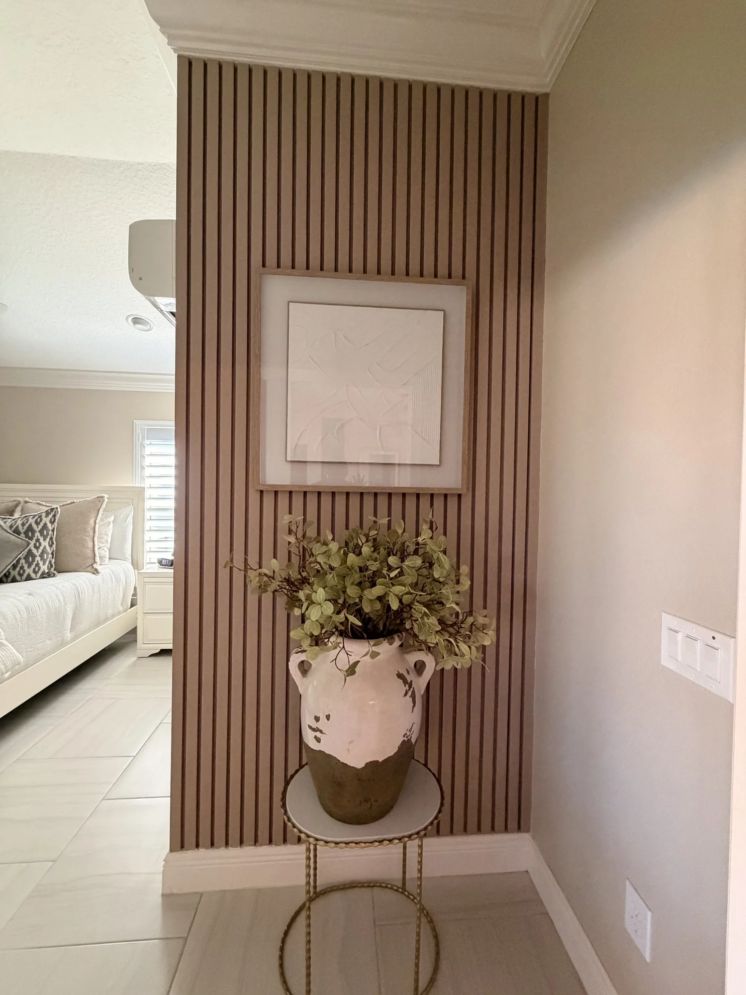 A decorative vase with greenery on a stand, placed against a wooden slat wall panel in a living room.
