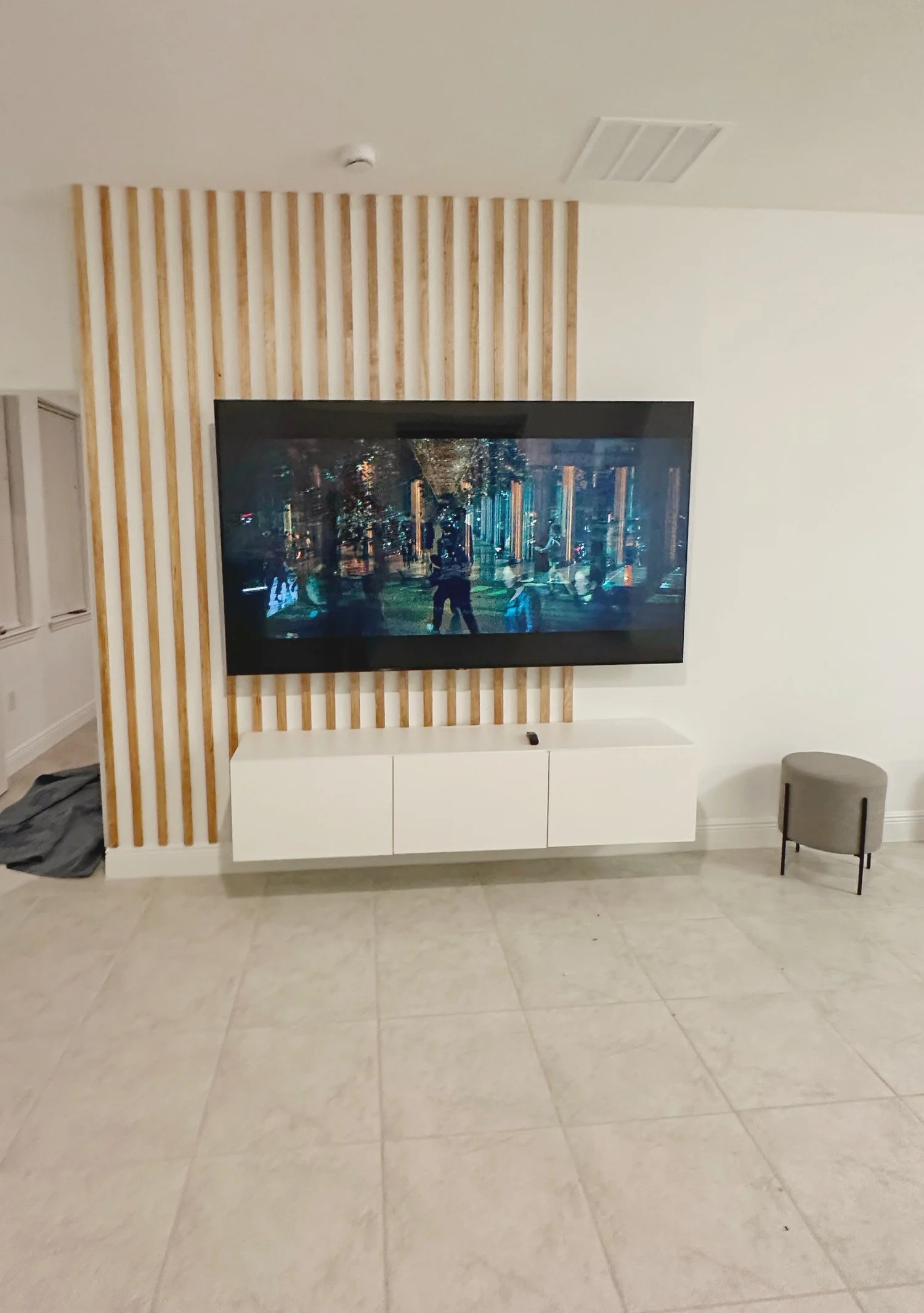 Living room with a large flat-screen TV mounted on the wall, a white media console below, a small gray ottoman to the right, and a wooden slat decorative wall behind the TV.