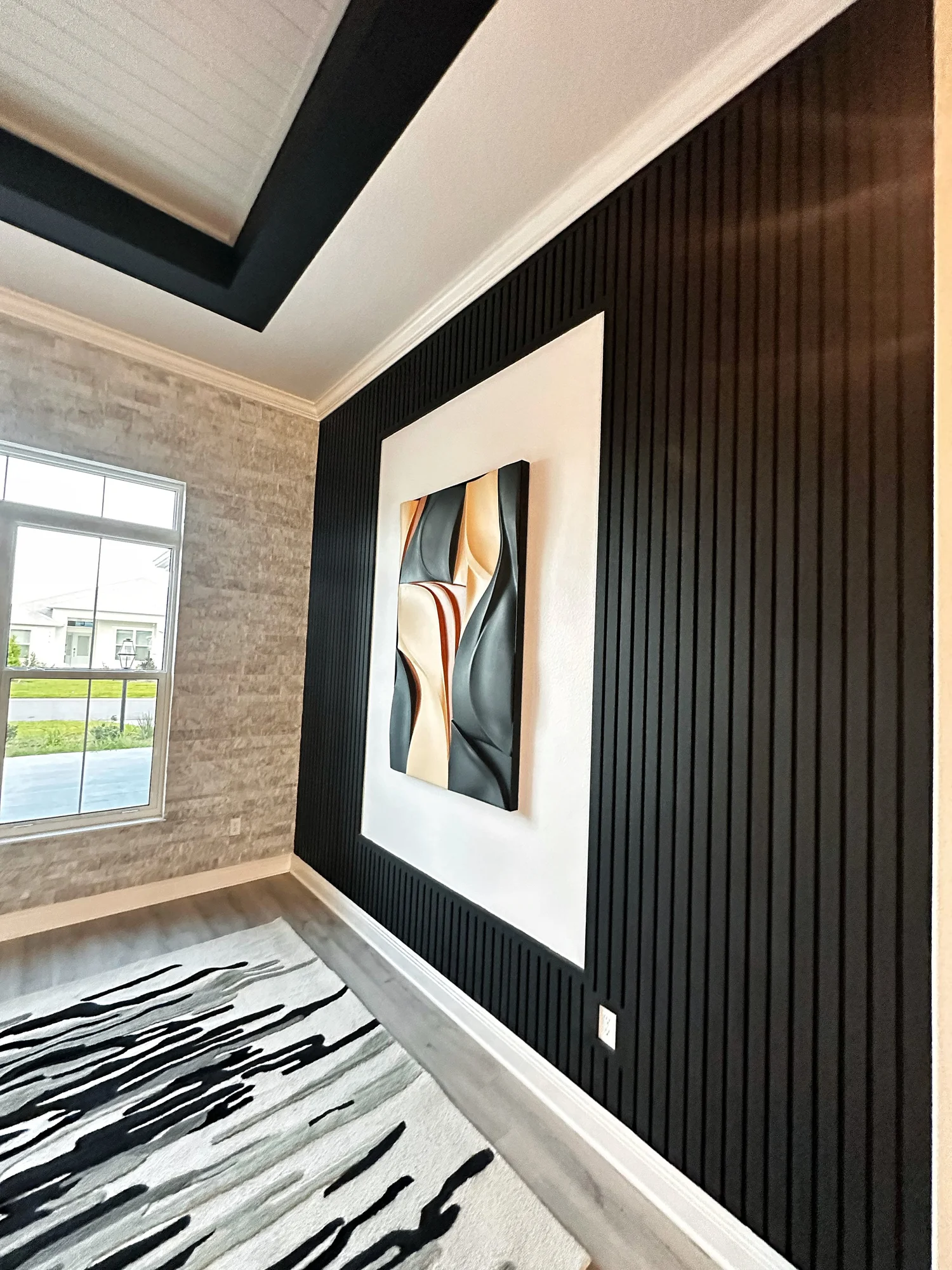 Interior of a modern living room with a black ribbed accent wall, a large white-framed abstract painting, a window with a view of a lawn and neighboring houses, and a black and white patterned rug.