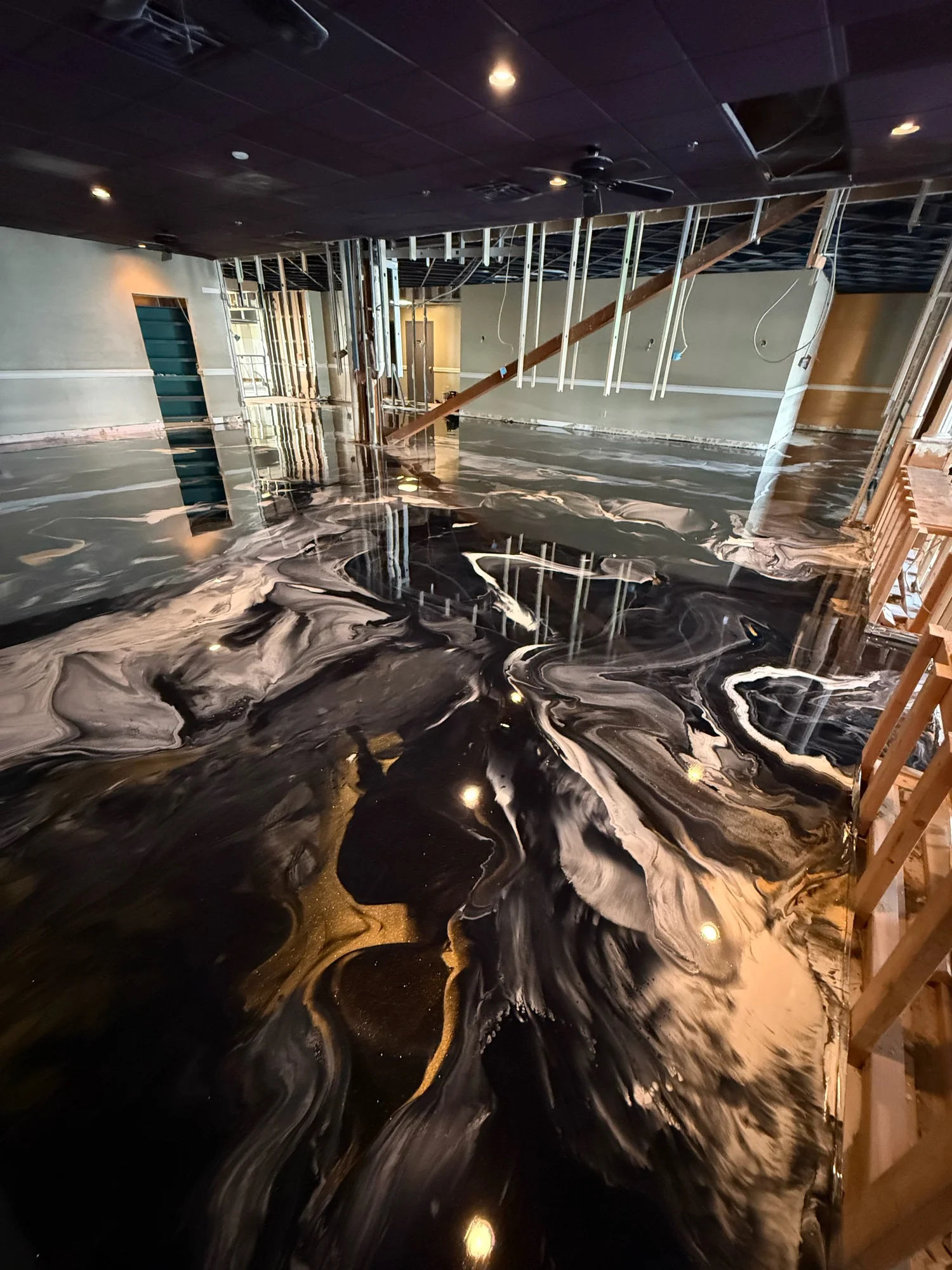 Interior of a building under renovation with a marble floor featuring black and white swirl patterns, unfinished walls, exposed wooden framing, and ceiling with recessed lighting and a black ceiling fan.