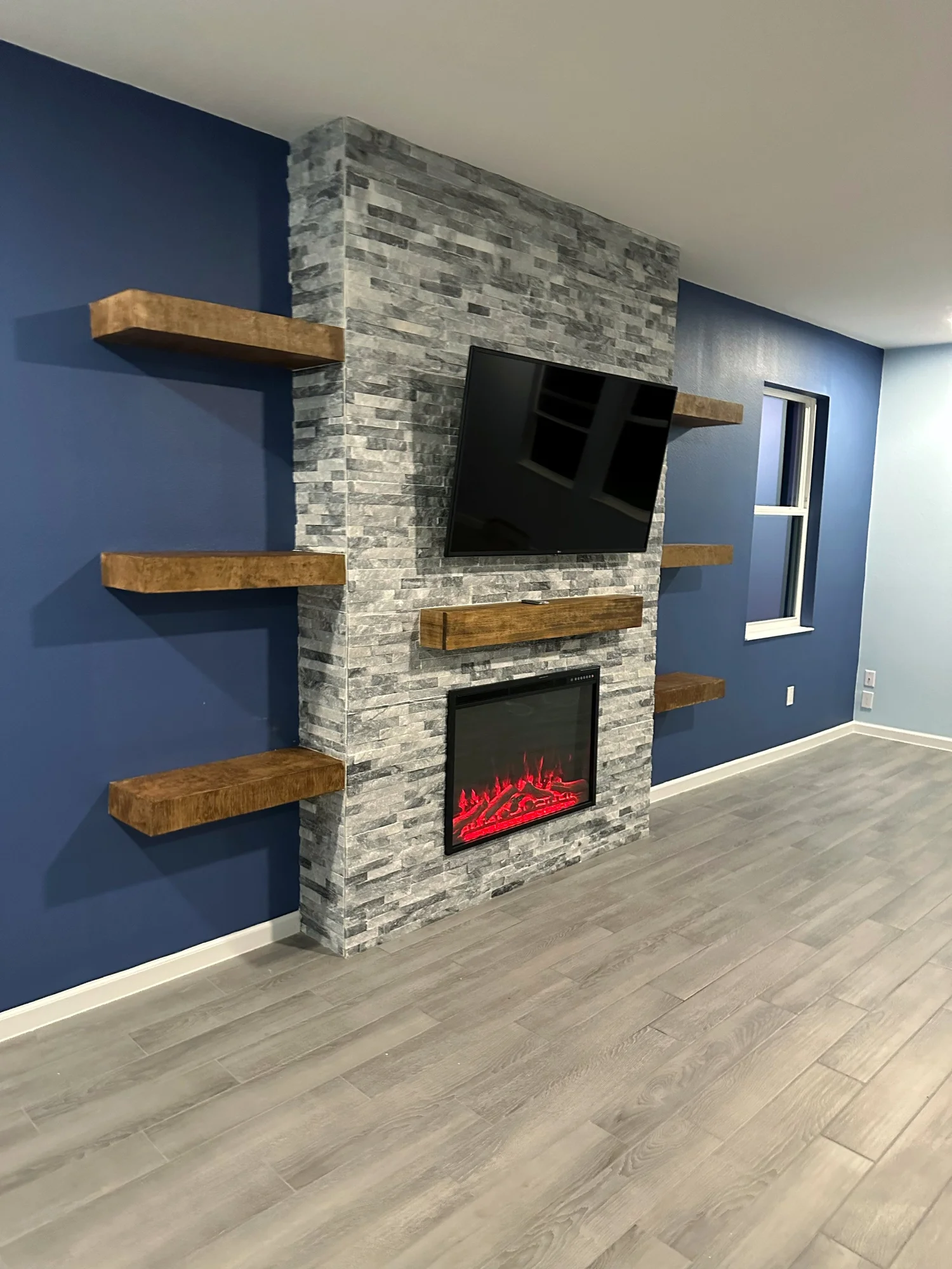 Living room with blue accent wall, stone fireplace with mounted TV, three wooden shelves, and gray wood flooring.