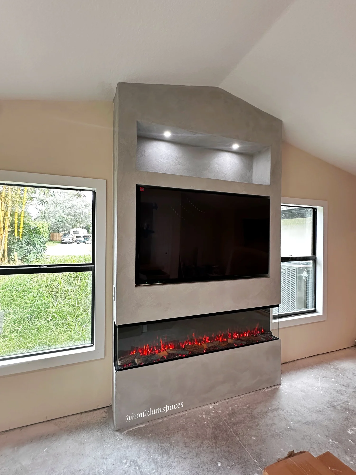 Modern living room fireplace with a mounted flat-screen TV above, and a rectangular electric fireplace below, set into a textured gray wall with spotlights, with two windows on either side showing outdoor greenery.