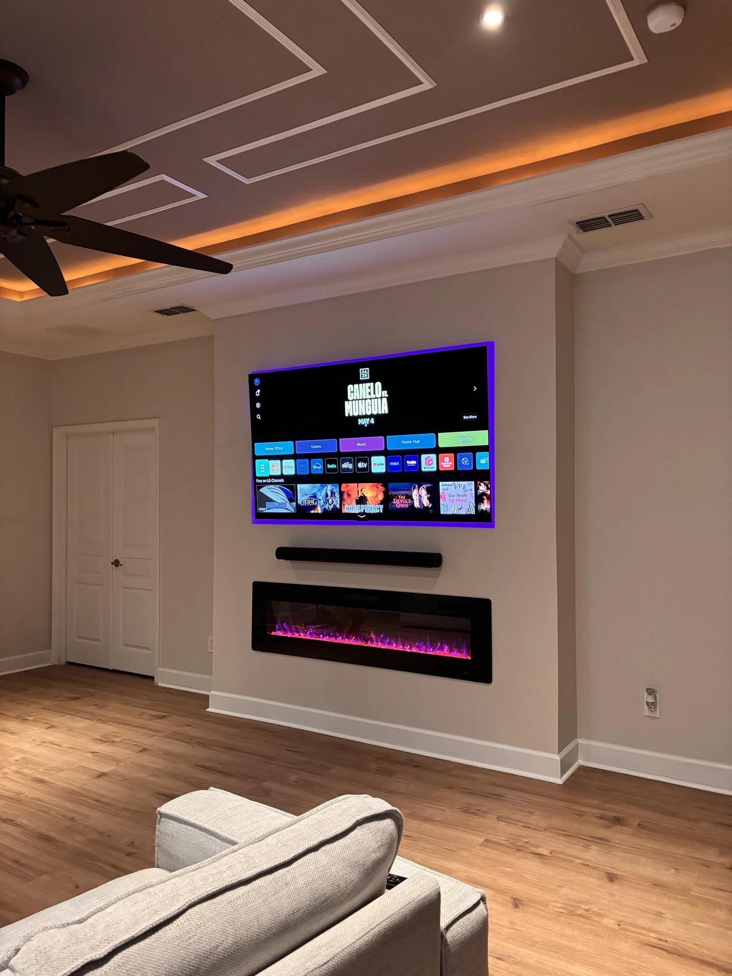 Modern living room with a wall-mounted flat-screen TV, a black electric fireplace beneath it, and a cozy beige armchair in the foreground. The ceiling features recessed lighting and decorative molding, with a ceiling fan nearby.