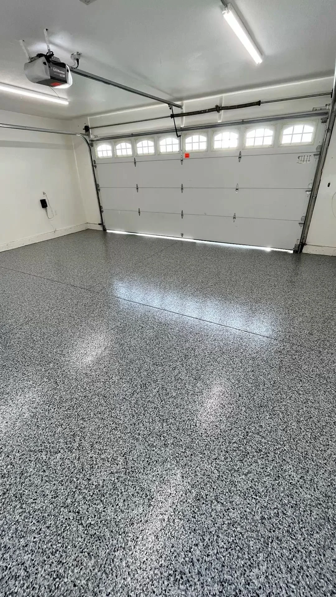 Empty garage with white walls, a speckled epoxy floor, and a closed white garage door with windows at the top. There is a garage door opener and some wiring on the ceiling.