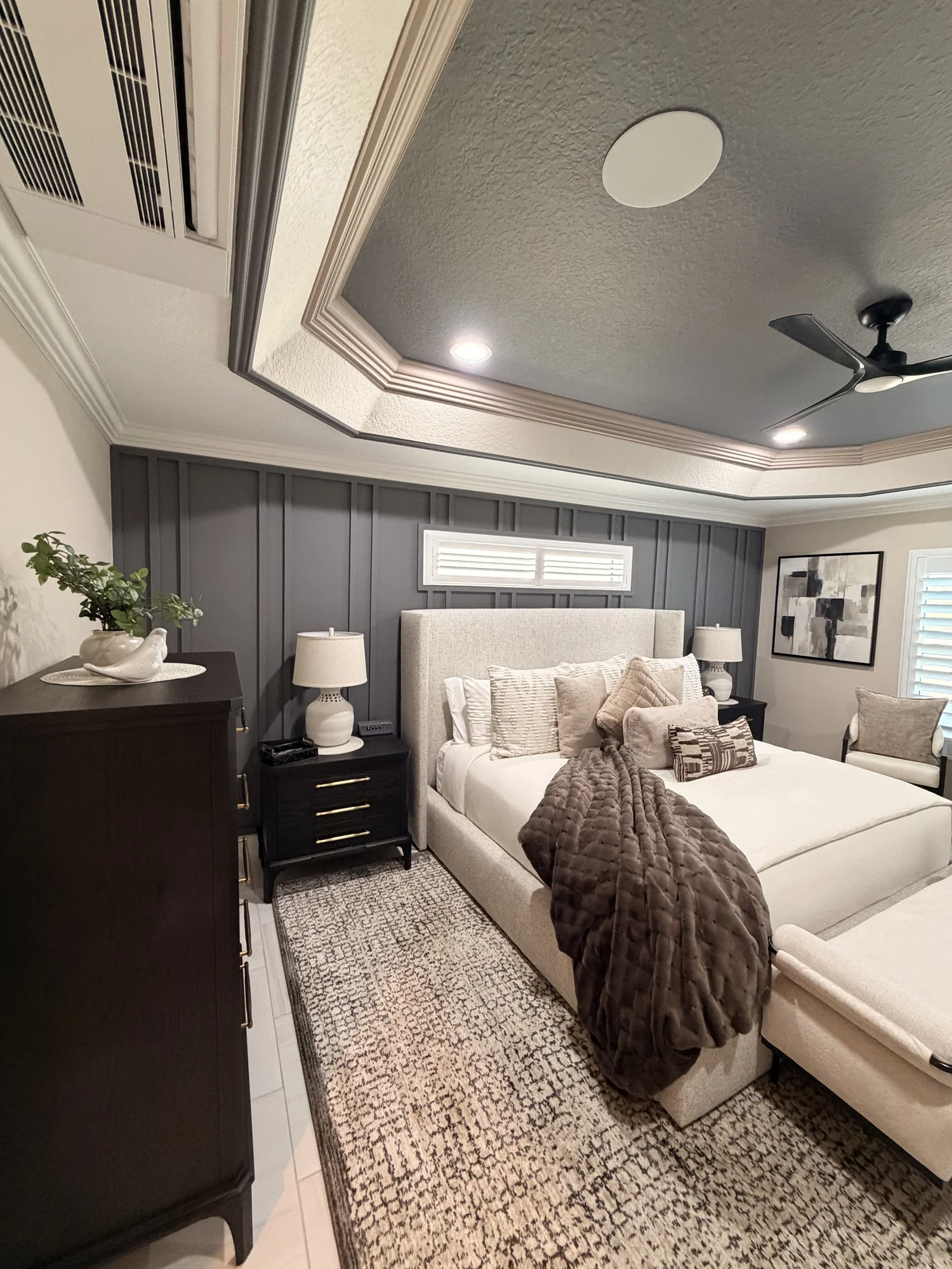 A bedroom with a beige upholstered bed, dark nightstands with white lamps, a patterned rug, and dark paneled walls. There is a window with white shutters, a framed abstract art piece on the wall, and a ceiling fan with integrated lighting.