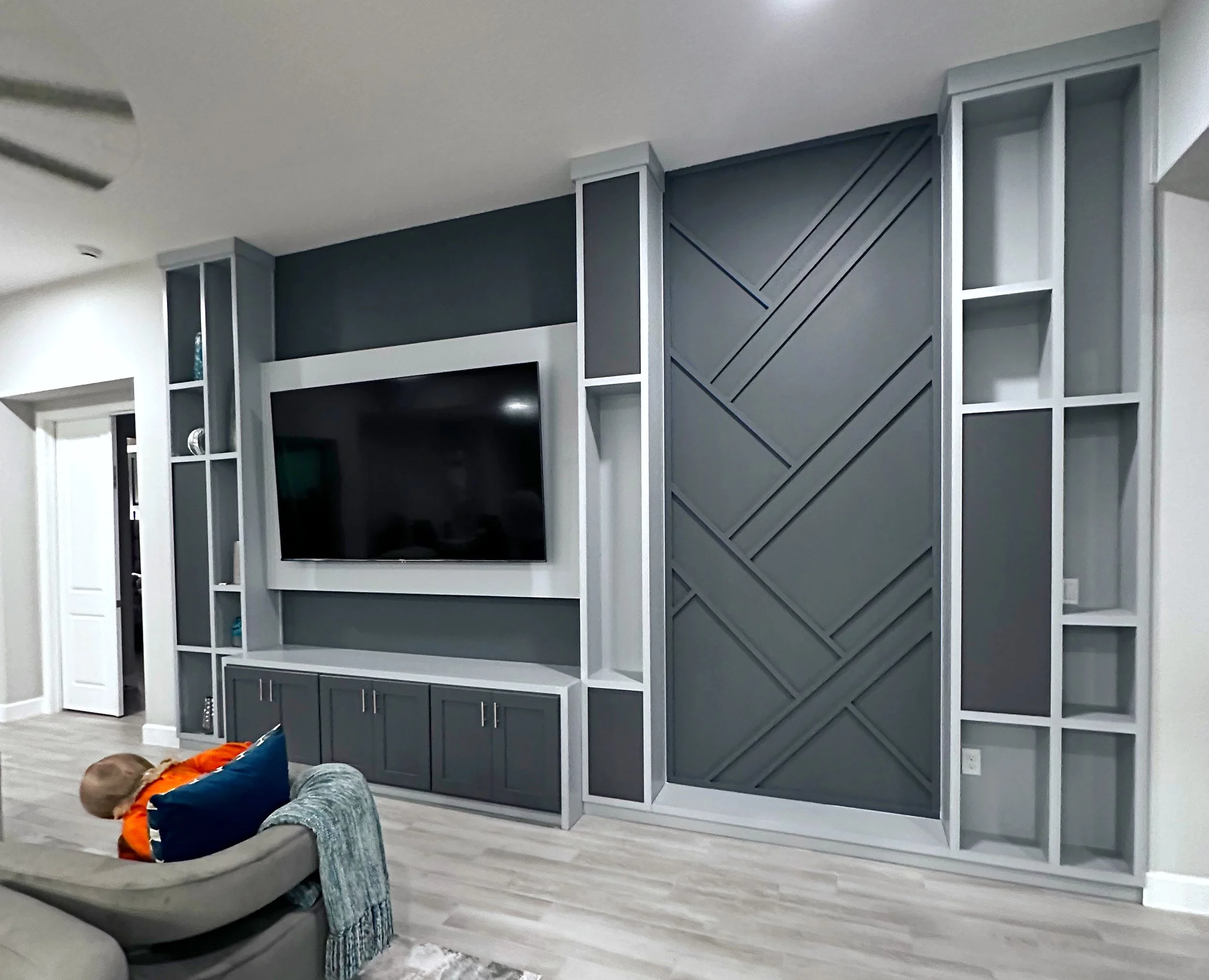 Living room with a built-in wall unit surrounding a large flat-screen TV, featuring geometric panel design in dark gray, with open white shelving on sides, light wood flooring, and a beige couch with a child resting on it.