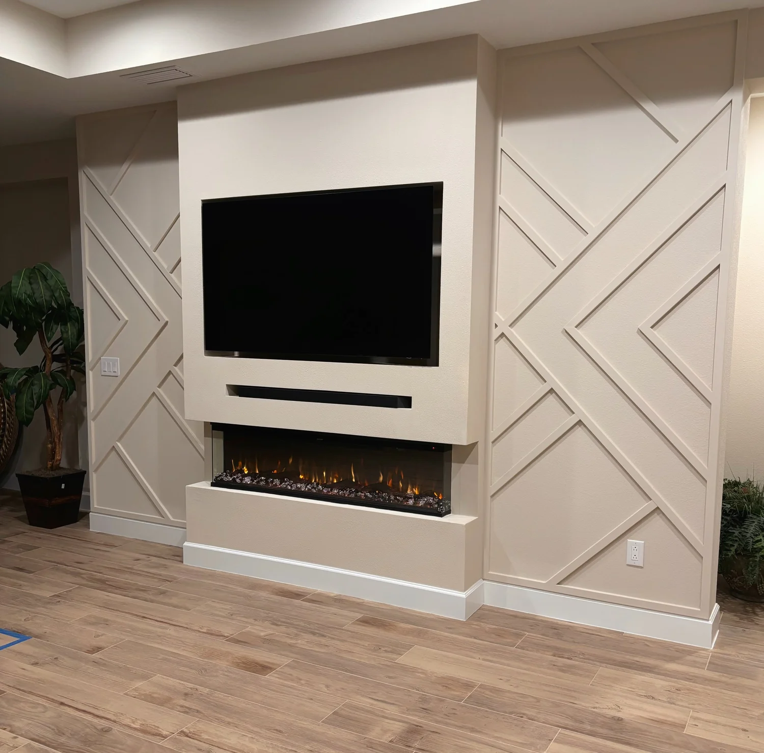Living room corner with a wall-mounted flat-screen TV above an electric fireplace, decorative wall panels with diagonal lines, hardwood flooring, and a potted plant in the background.