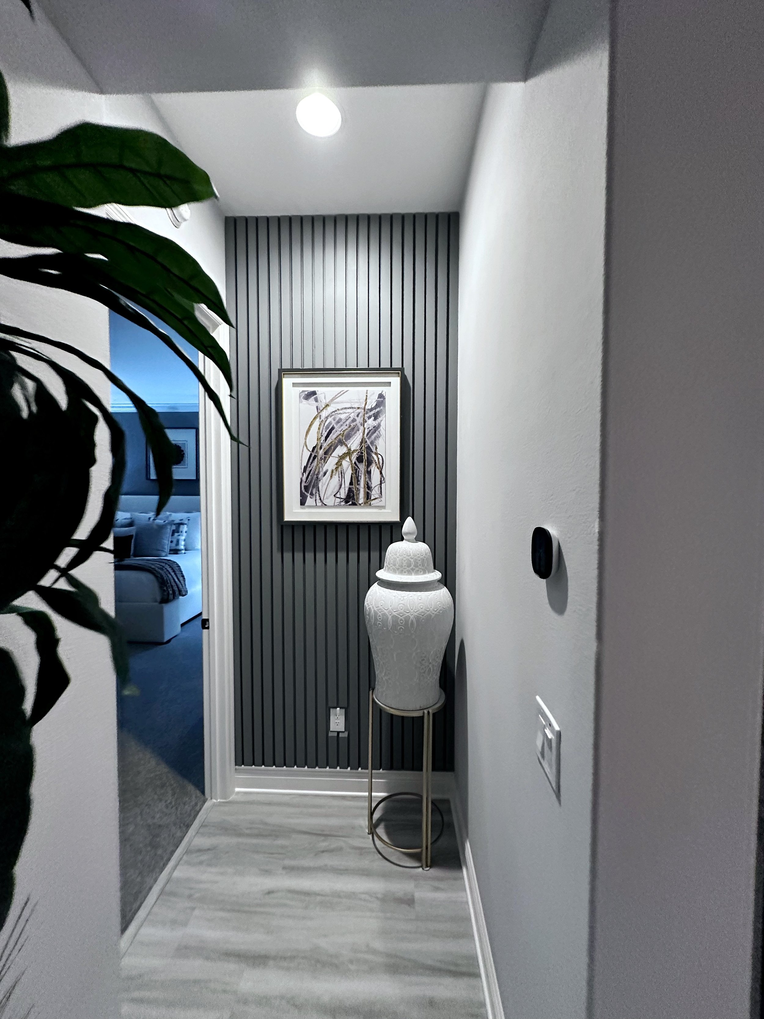 Hallway corner with black vertical striped wall, white decorative vase on a stand, framed abstract art above, and a ceiling light.