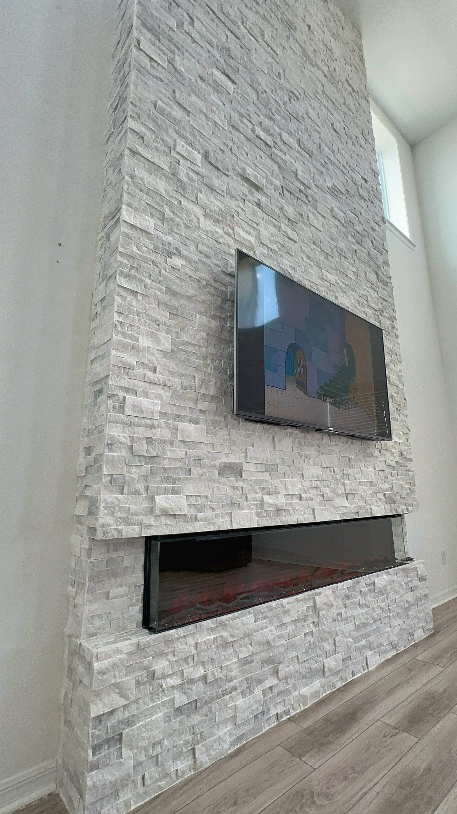 A modern interior fireplace wall made of white stone with a TV mounted above and a glass-enclosed electric fireplace below, in a bright room with high windows.