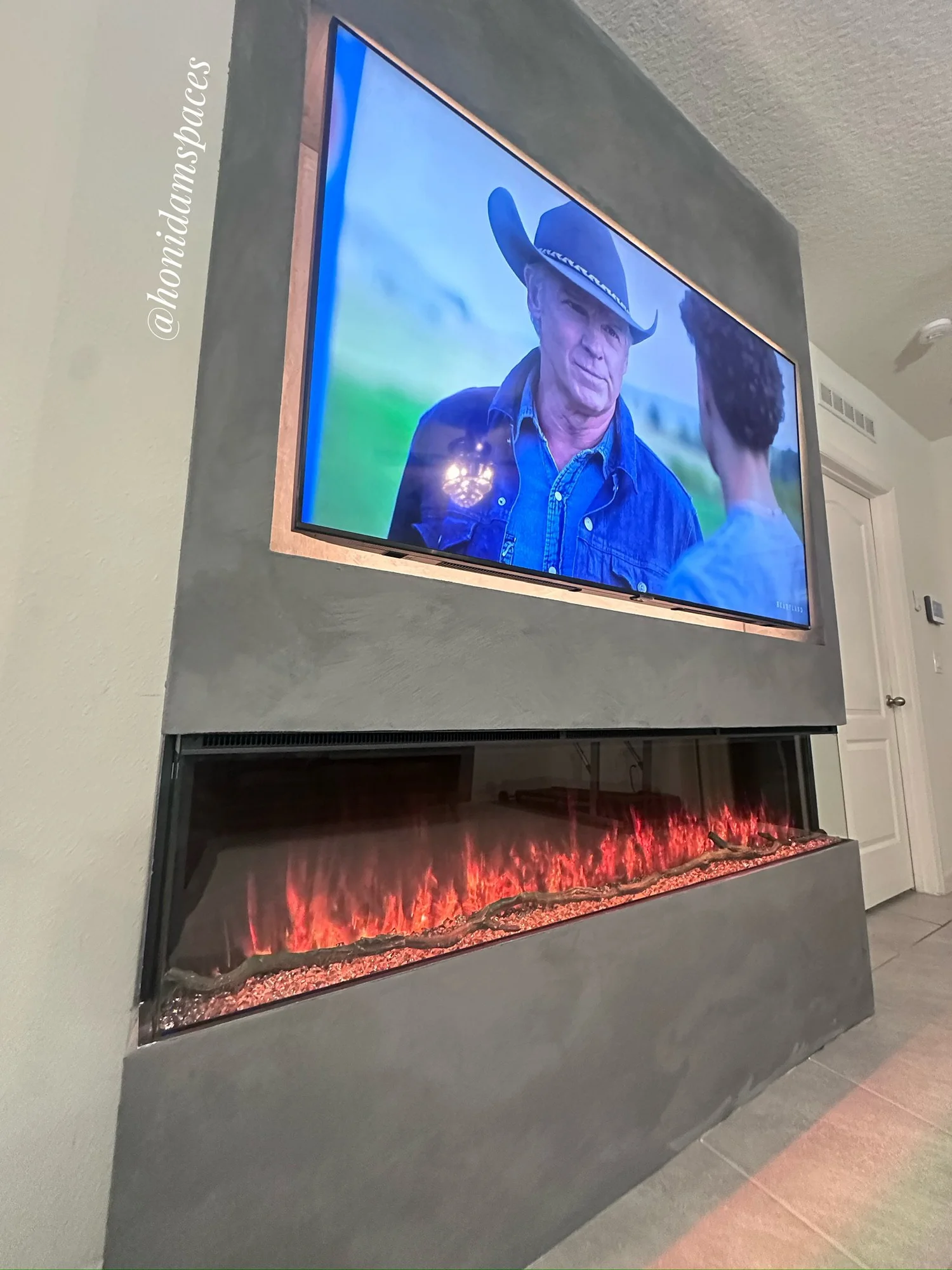Wall-mounted flat-screen TV displaying a scene with a man in a cowboy hat and denim jacket talking to a woman, above an electric fireplace with red flames and decorative branches inside.