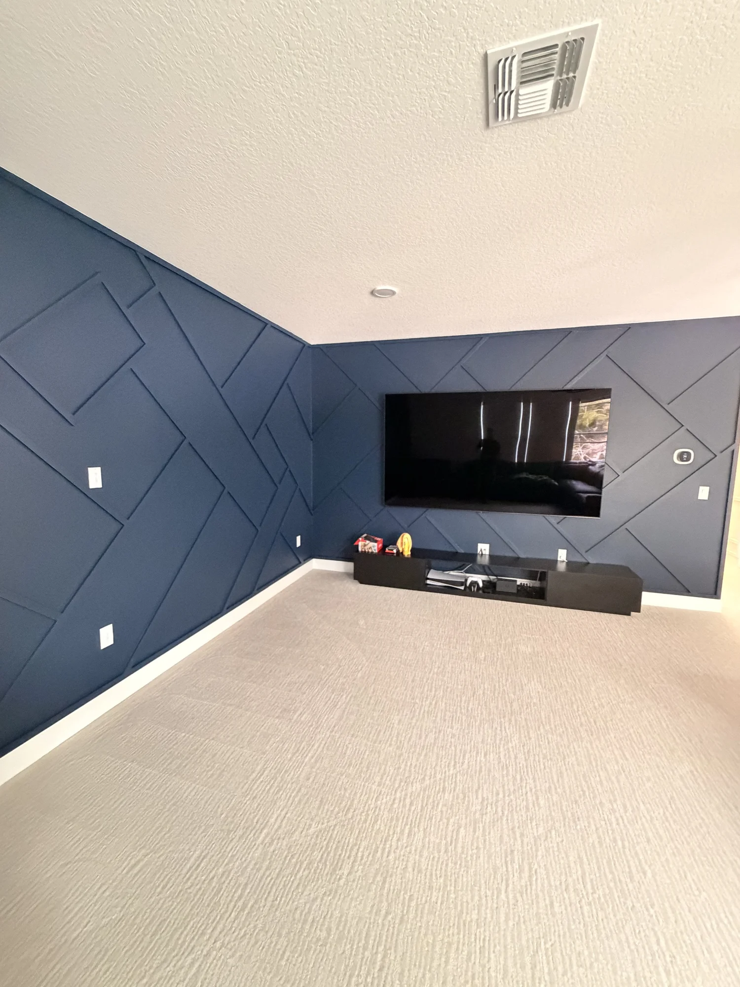 A living room corner with a dark blue textured accent wall, a mounted flat-screen TV, and a black entertainment console with a few decorative items on top. The room has a beige carpet and a textured white ceiling with a vent.
