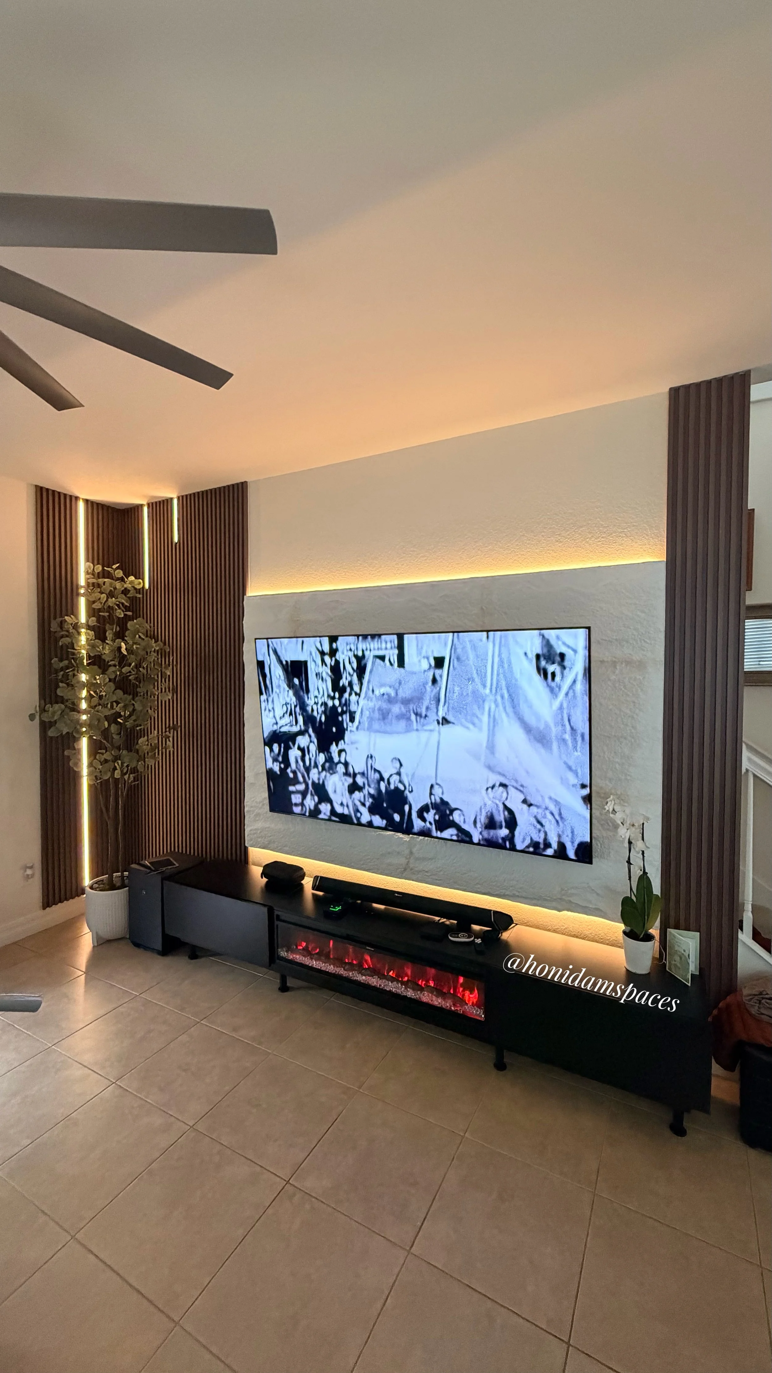 Living room with a wall-mounted TV, a black TV stand, a fireplace with red flames, a potted plant, and wooden decorative panels on the walls.