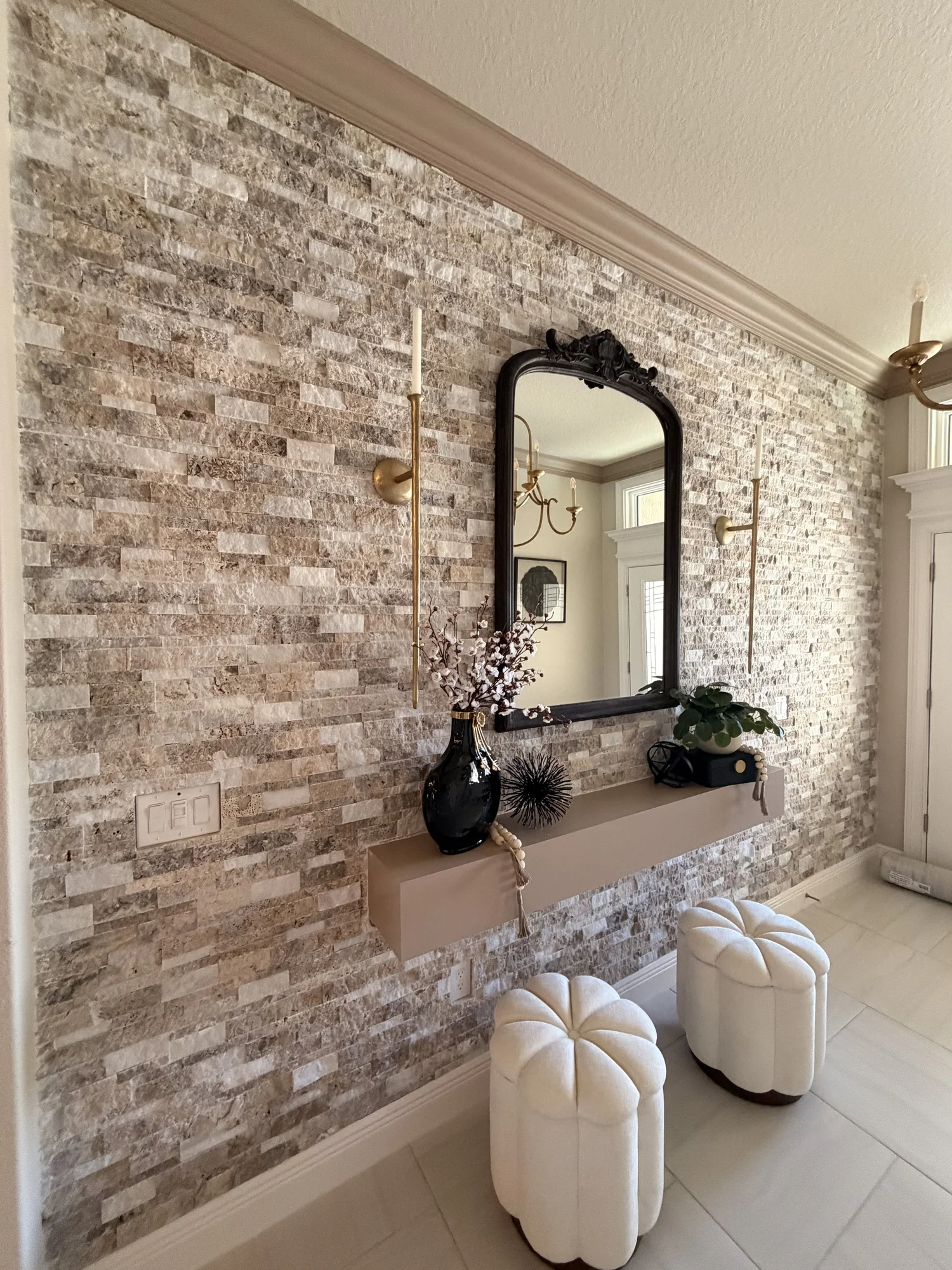 Interior wall with brick stone veneer, a large black framed mirror, a decorative black vase with white flowers, a black box, and two white tufted ottomans.