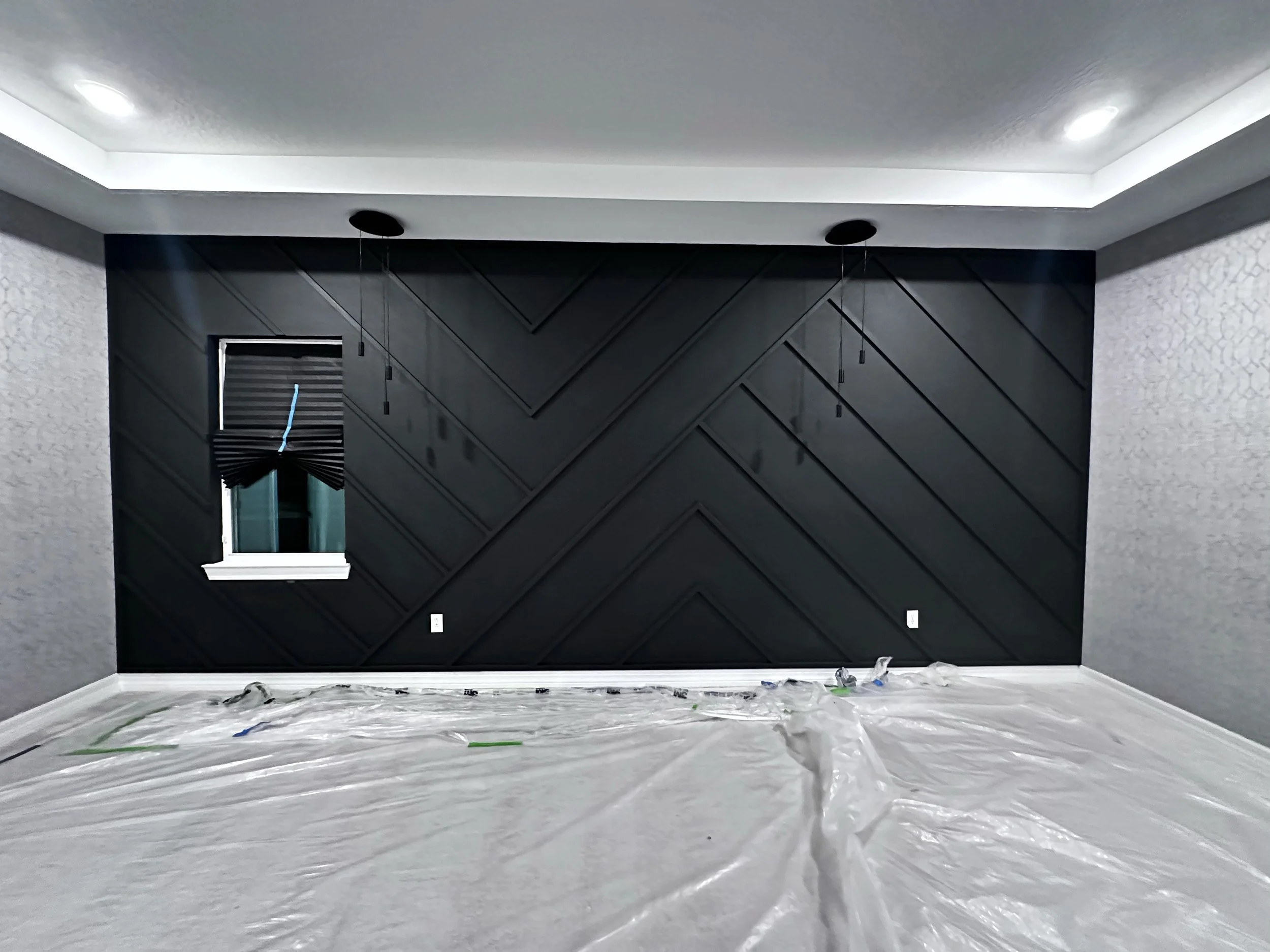 Room with black accent wall featuring geometric paneling, window with blackout shade, and plastic covering on the white floor