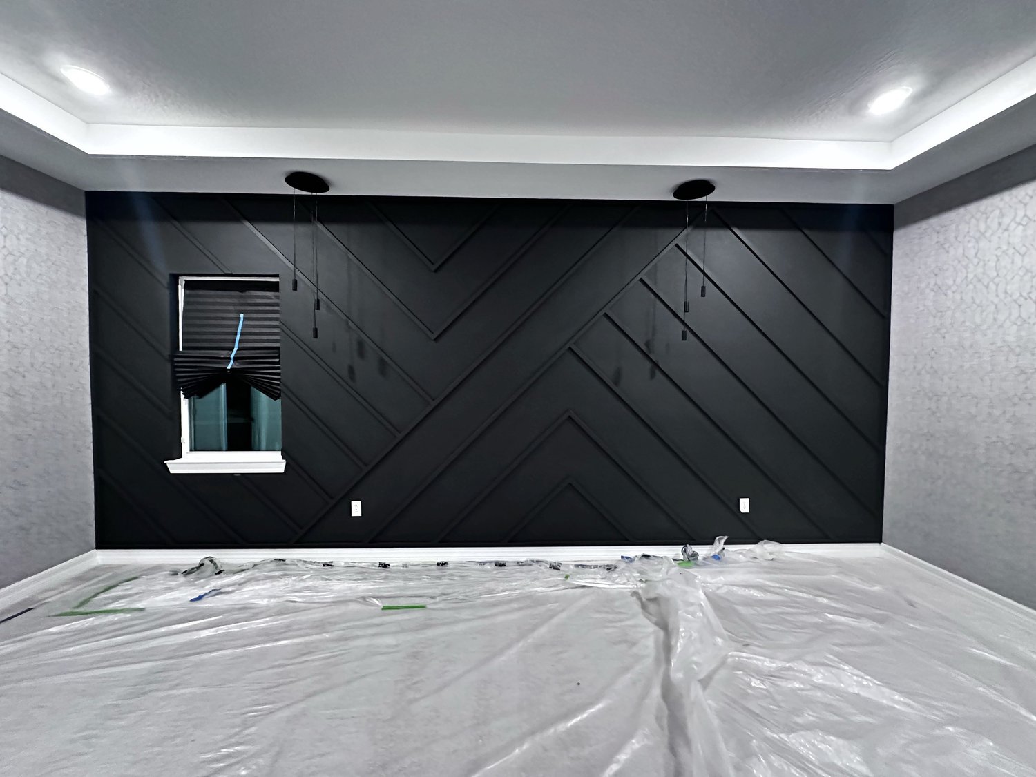 Room with black accent wall featuring geometric paneling, window with blackout shade, and plastic covering on the white floor