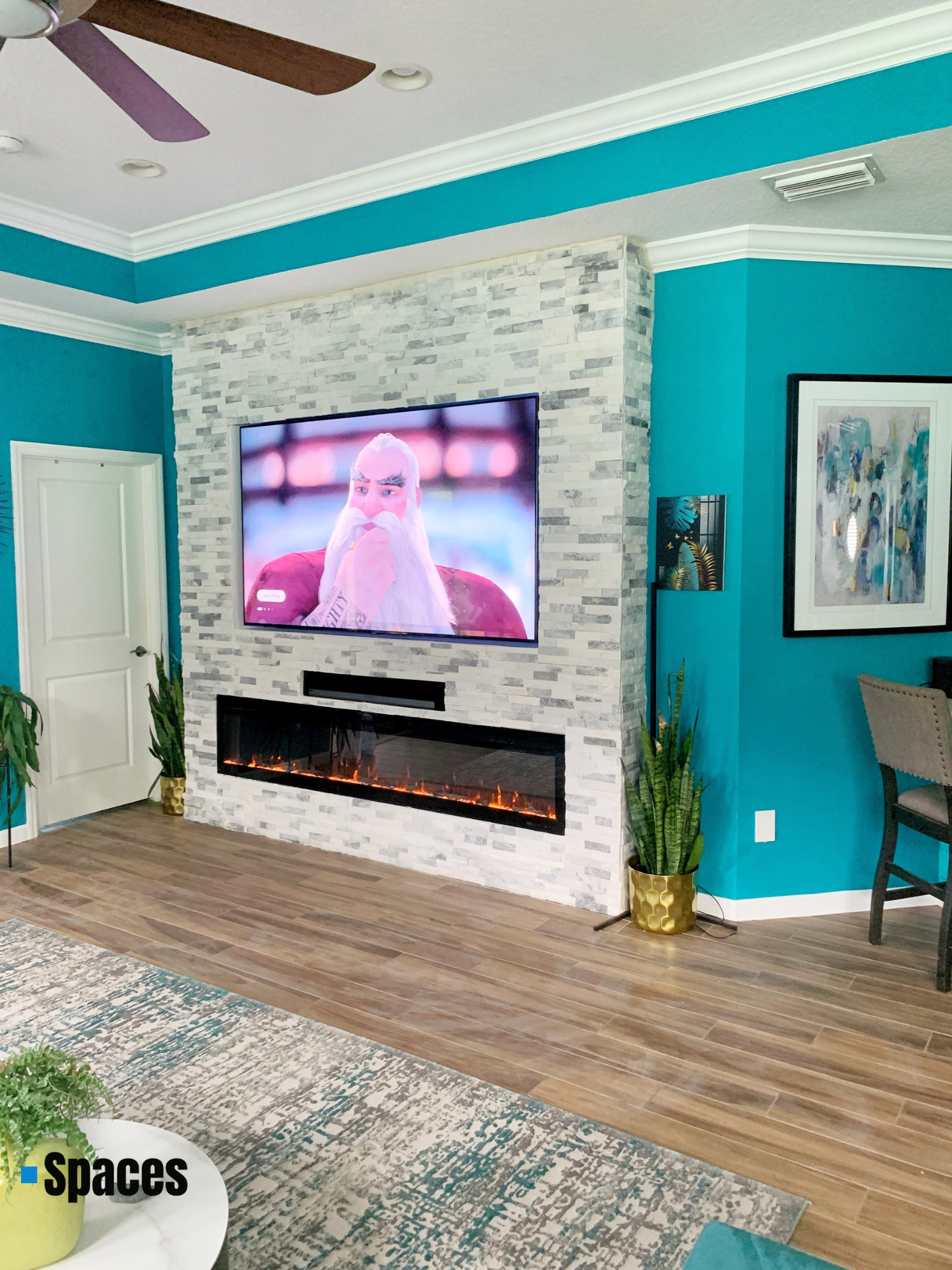 Living room with turquoise walls, a wall-mounted TV above a modern electric fireplace with a white and gray stone surround, potted plants, a framed colorful abstract painting, a chair, and a ceiling fan.