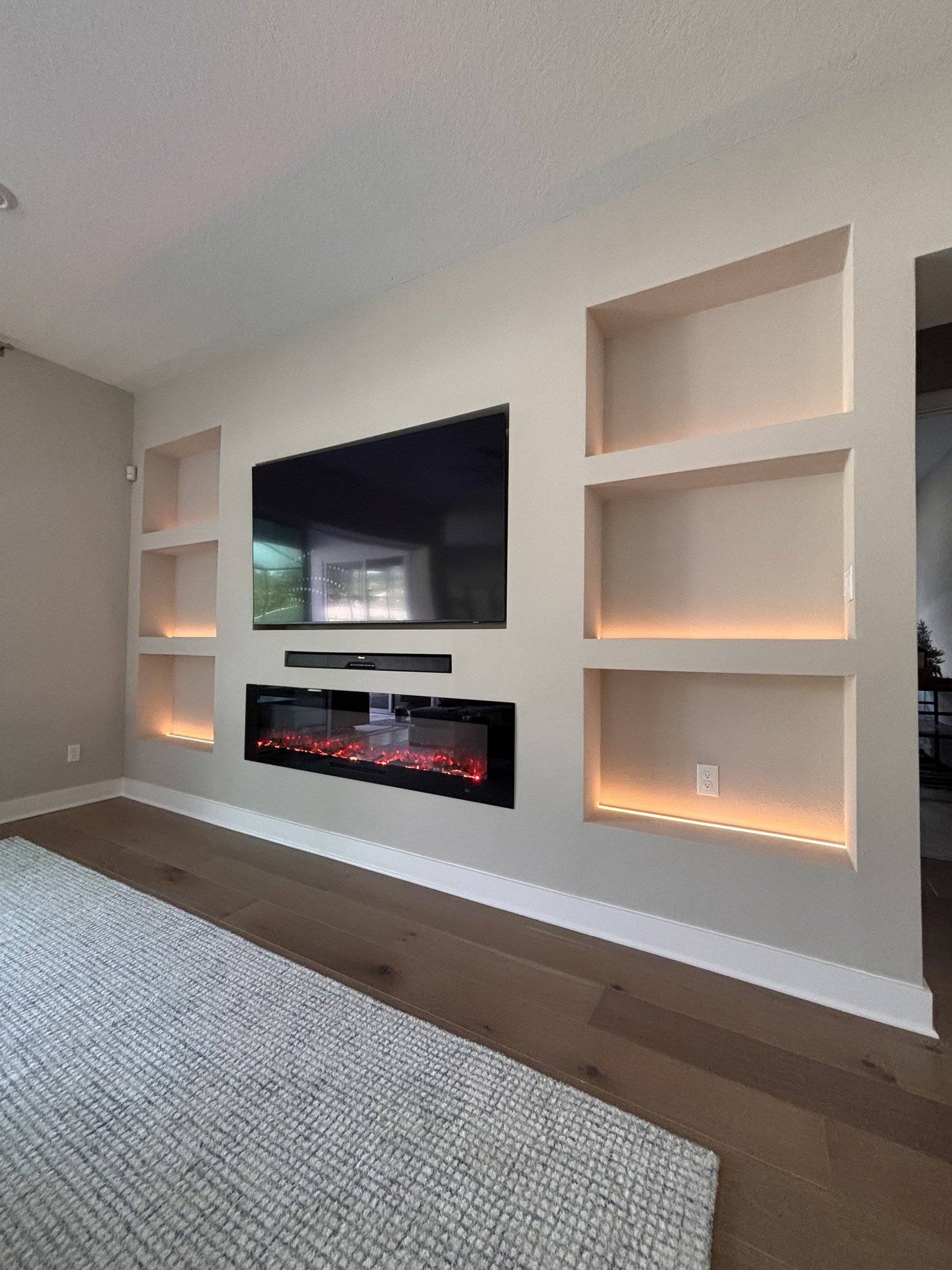 Modern living room wall with a mounted flat-screen TV, an electric fireplace below it, and three built-in wall shelves with ambient lighting.
