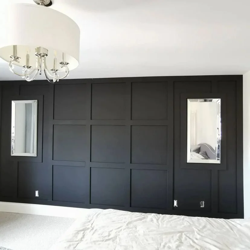 A bedroom wall with black paneled wainscoting, two narrow rectangular mirrors, and a white ceiling with a chandelier. Part of a bed with white bedding is visible in the foreground.