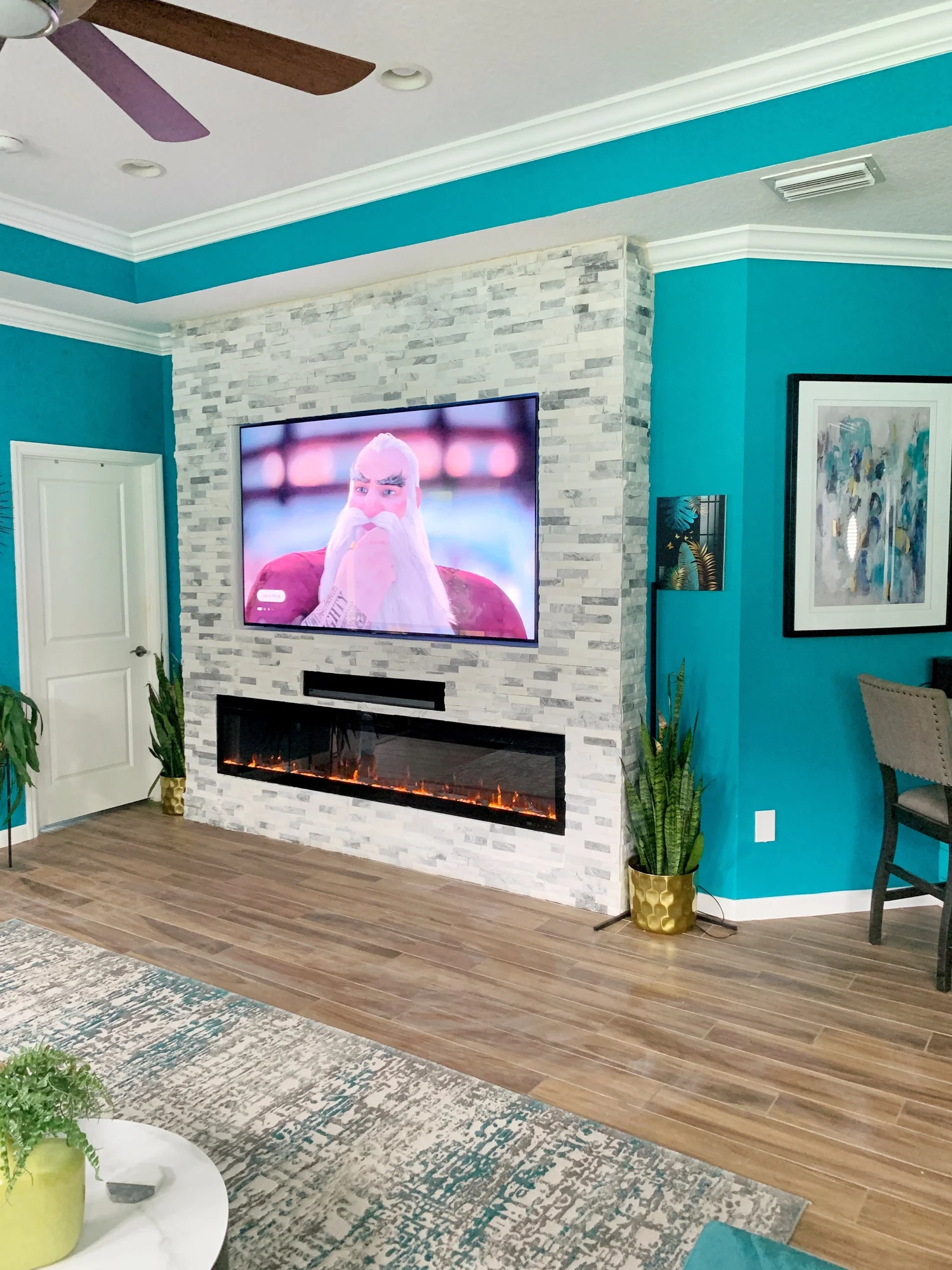Living room with a mounted flat-screen TV above a modern electric fireplace, surrounded by white and gray stone, turquoise walls, and decorated with potted plants, a colorful area rug, and framed artwork.