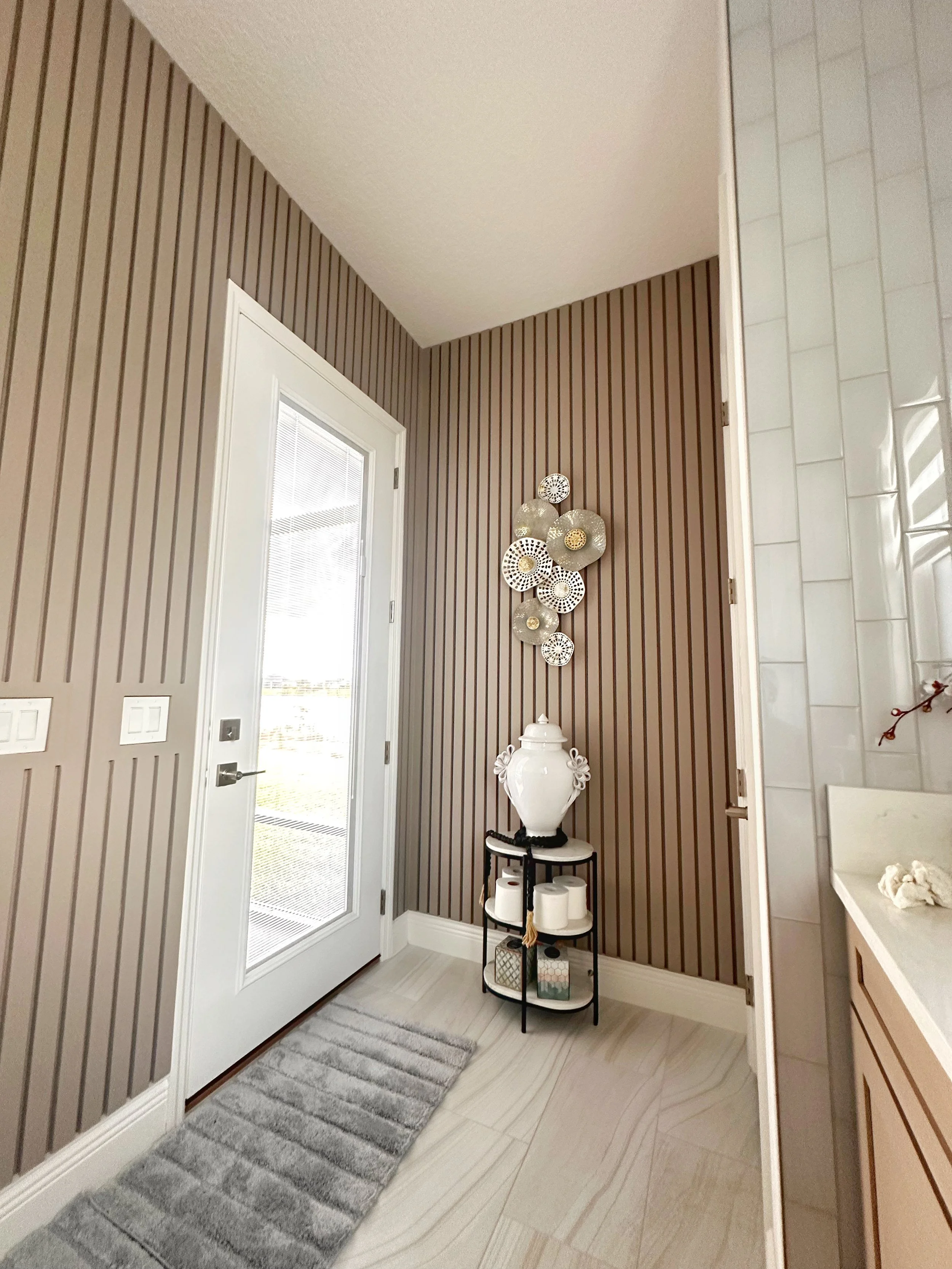 A corner of a bathroom with a door, decorative wall art, a lamp, and a small table holding a vase and toilet paper rolls.