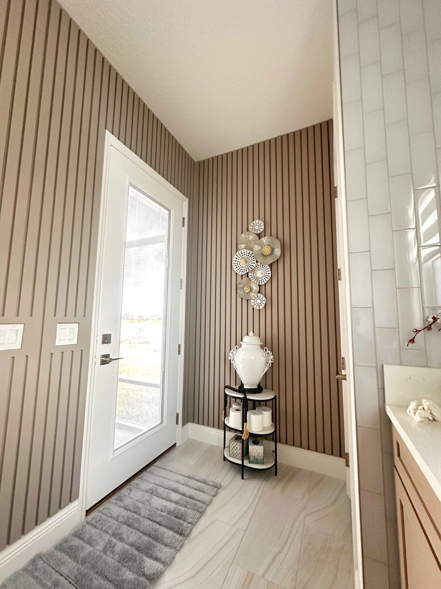 A corner of a bathroom with a door, decorative wall art, a lamp, and a small table holding a vase and toilet paper rolls.