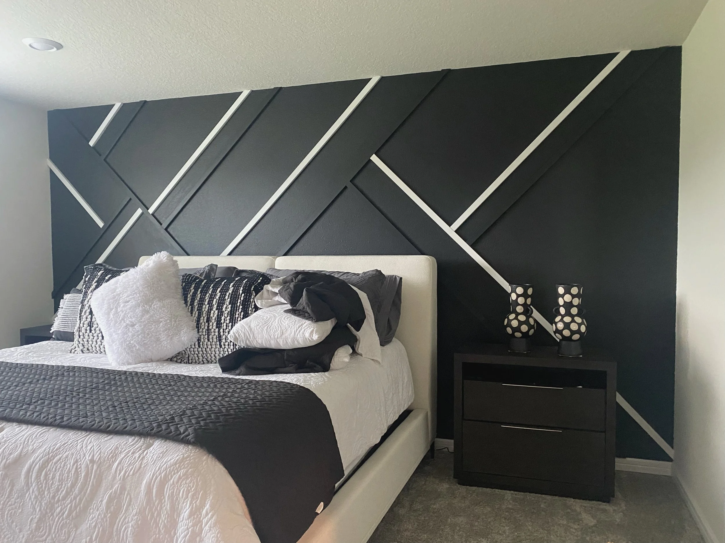Bedroom with a bed, pillows, and a black nightstand with two matching black and white polka dot vases against a dark geometric accent wall.
