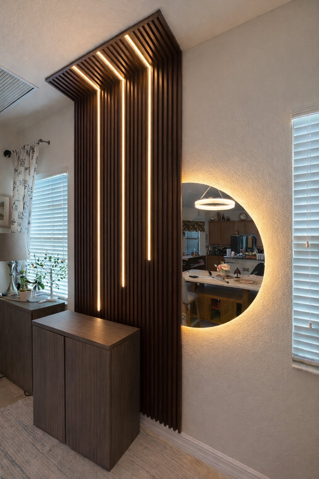 Interior of a home with a wooden slat room divider featuring vertical LED strip lights, and a circular mirror with backlighting on a textured wall, reflecting a dining area and kitchen.