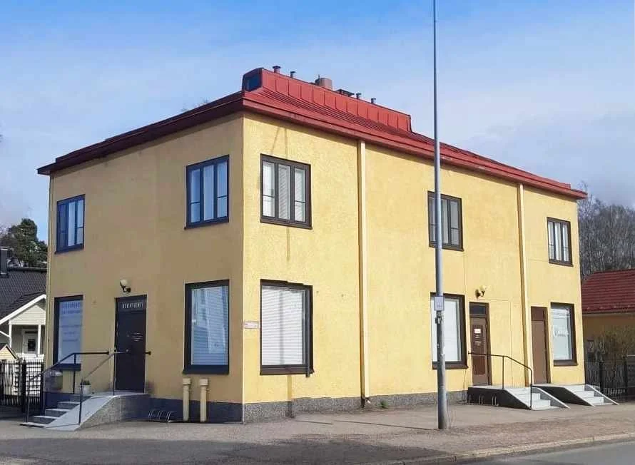 Yellow two-story building with black window frames, red sloped roof, and two entrance doors with stairs outside, situated on a paved street with a metal lamp post in front.