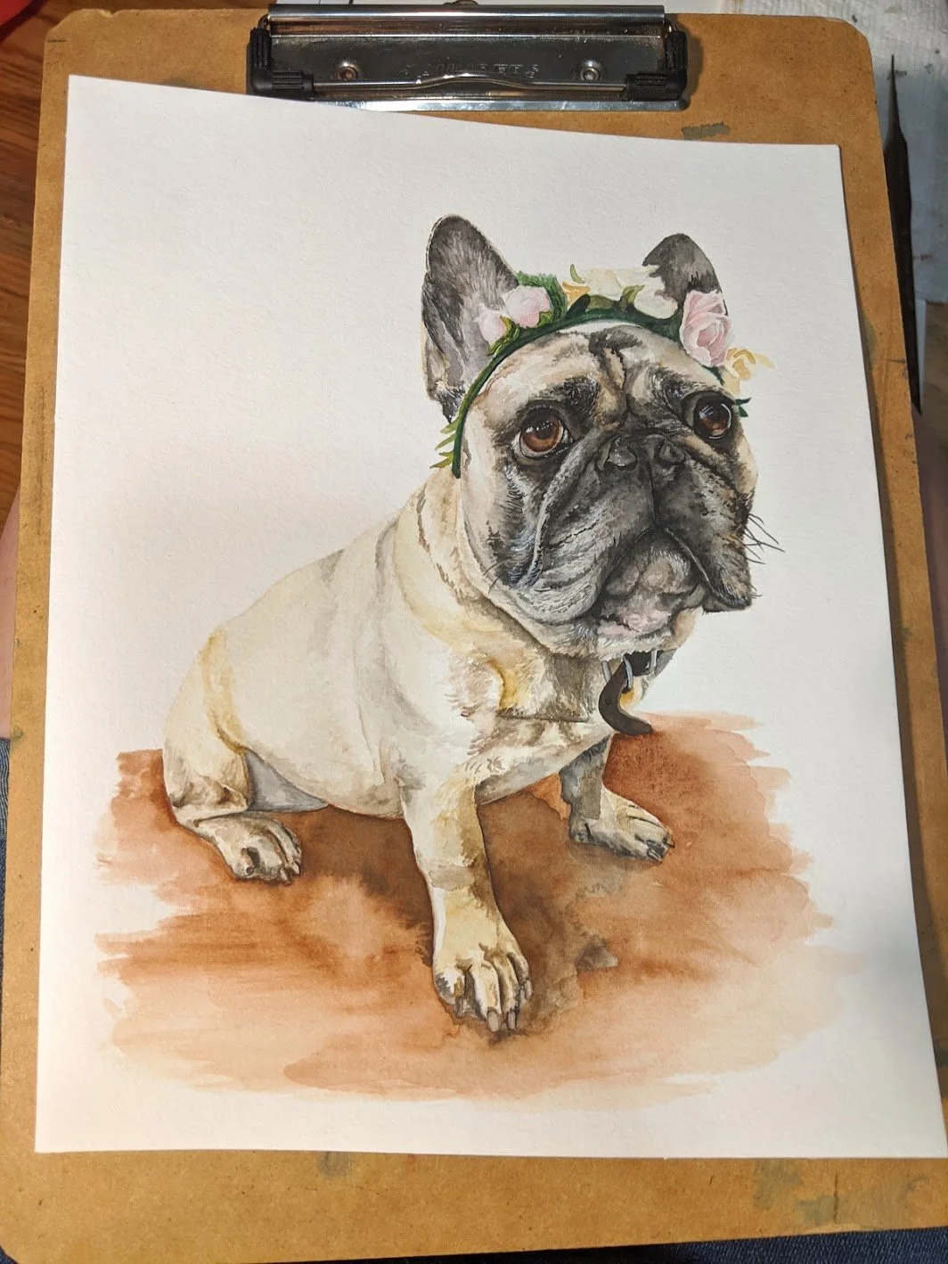 Photograph of a watercolor painting of a French Bulldog with a flower crown on its head, sitting on a wooden surface.