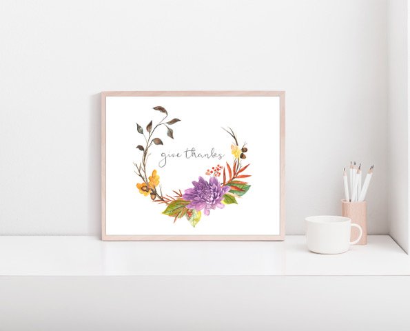 Decorative framed art with the words "give thanks" surrounded by colorful watercolor flowers and leaves, placed on a white shelf next to a white mug and a pink container with pencils.