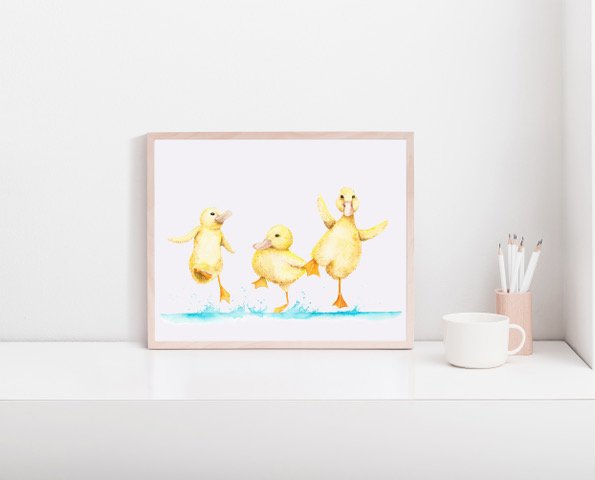 Framed artwork of three yellow rubber ducklings standing on a white surface with a blue water background, next to a white mug, pink container, and pencils.