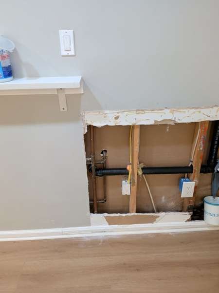 Opened wall section exposing plumbing behind removed drywall and baseboard