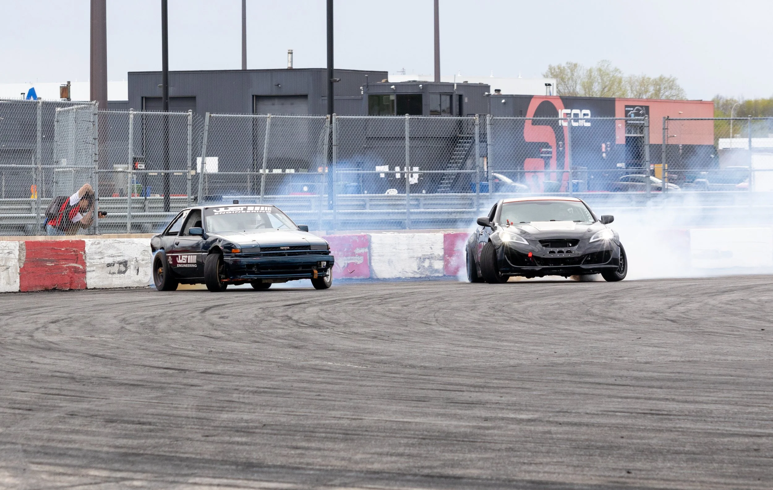 Two race cars drifting on a track, with smoke rising from the tires, behind a chain-link fence in a racetrack setting.