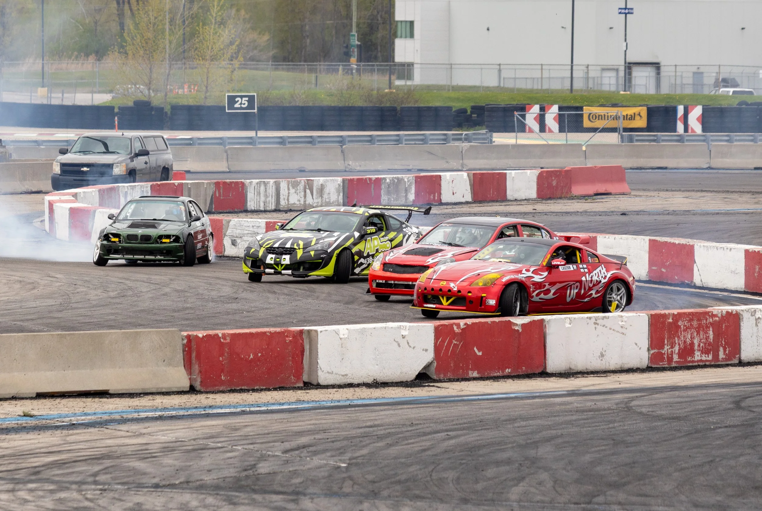 Race cars on a dirt track during a race, with barriers and a backdrop of trees and buildings.