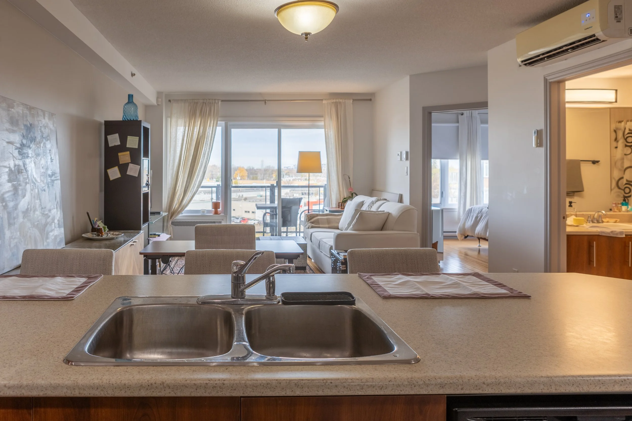 Living room with a white couch, a sliding glass door leading to a balcony, and a small bedroom visible through an open door, with a kitchen counter in the foreground and a sink.