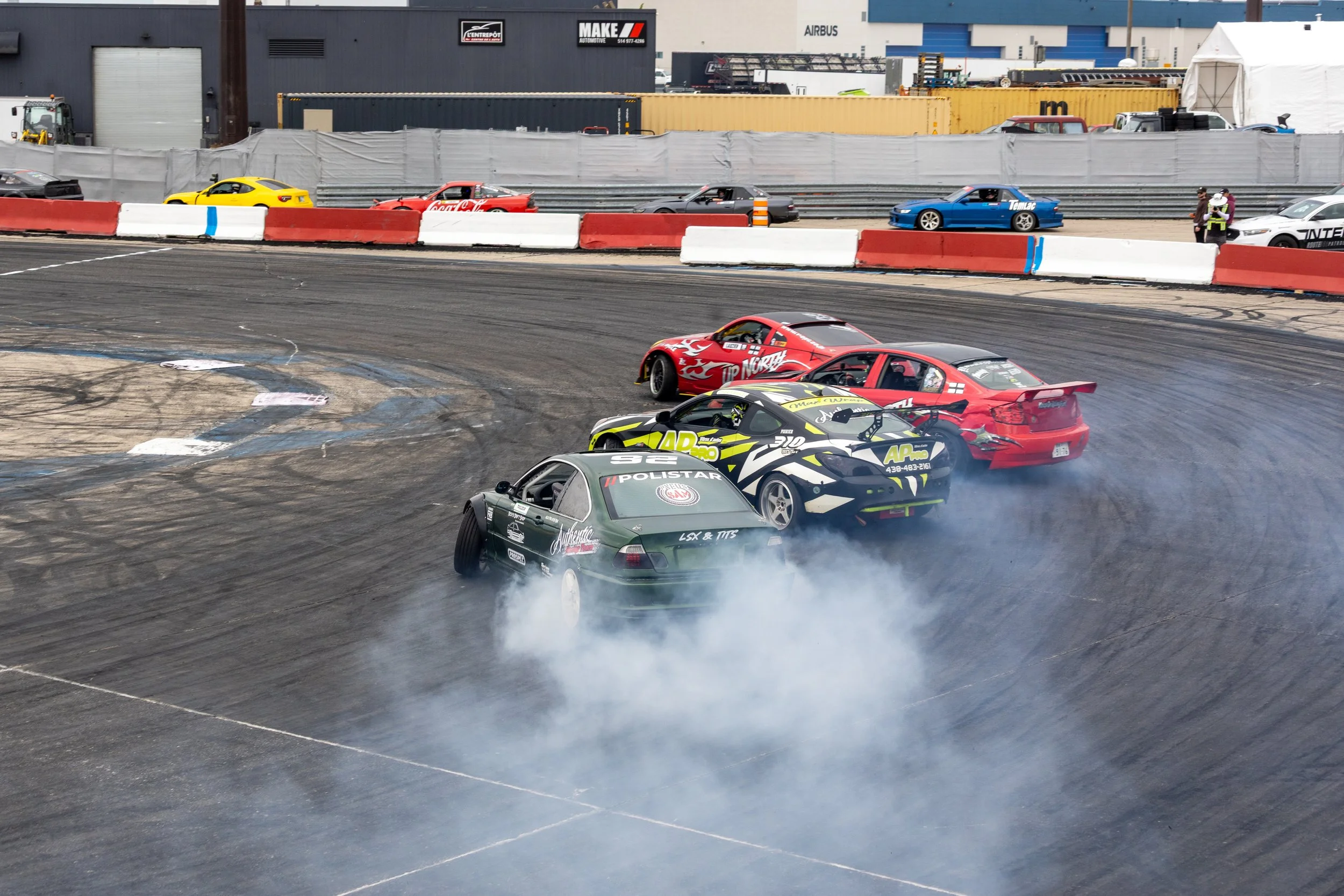 Race cars drifting around a corner on a racetrack, with tire smoke billowing behind them, and a backdrop of industrial buildings and advertisements.