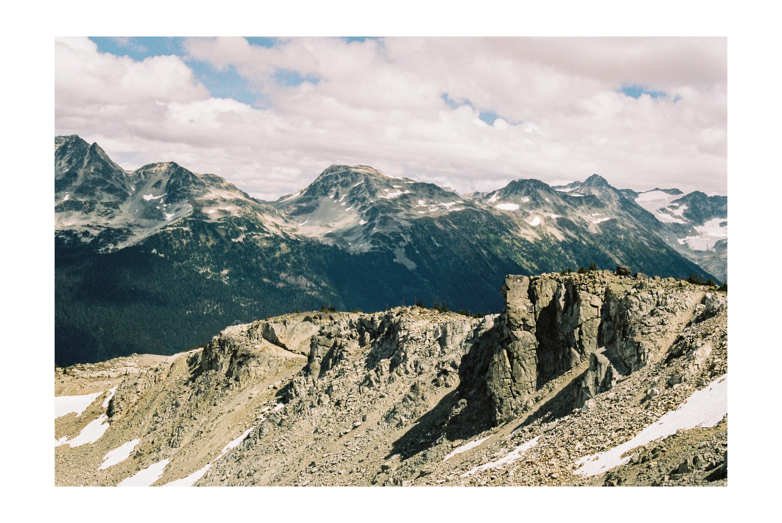 Mountain landscape with rugged rocks in the foreground, snow patches, and snow-capped peaks in the background under a partly cloudy sky. Whistler, BC. Shot on Kodak Pro Image 100.