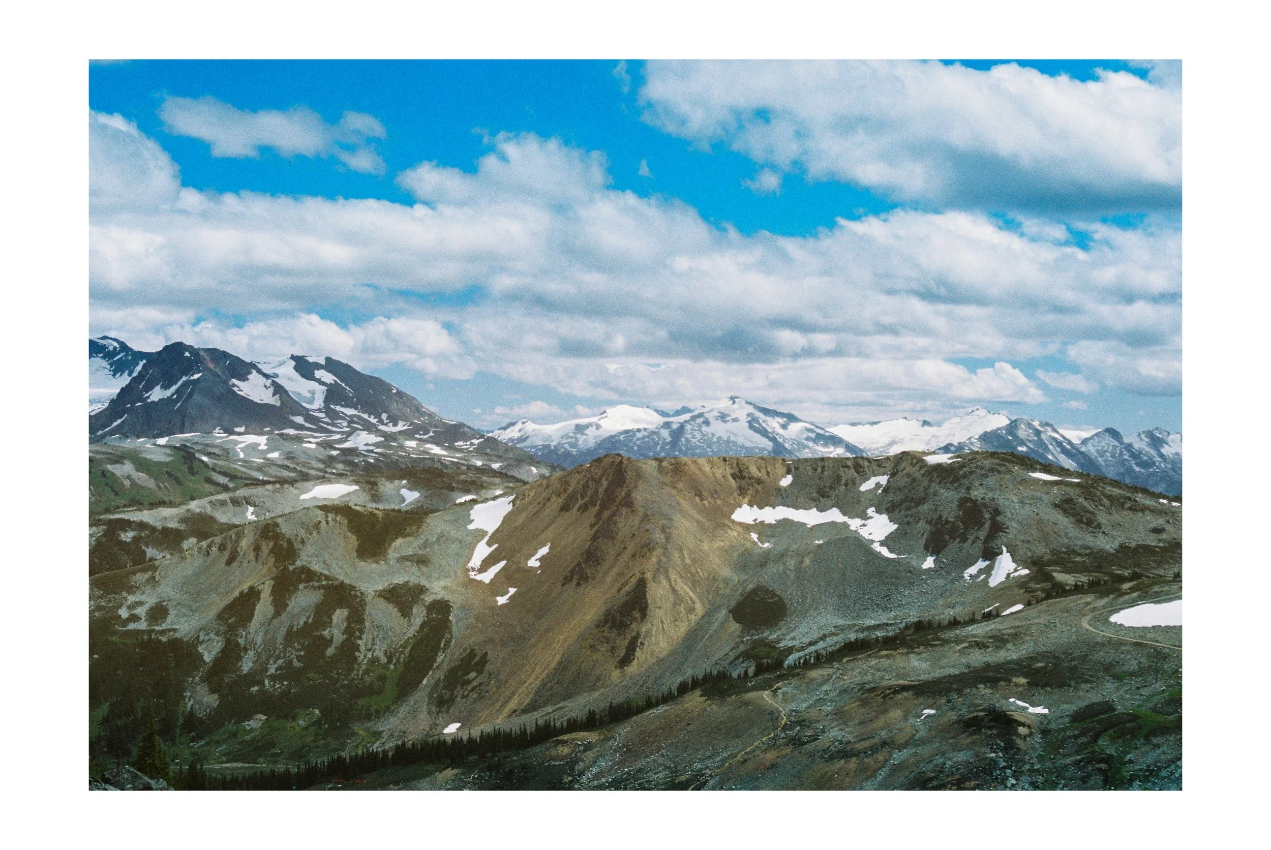 A mountainous landscape with snow-capped peaks, rugged terrain, and a partly cloudy blue sky. Taken on Kodak Pro Image 100 in Whistler, BC.