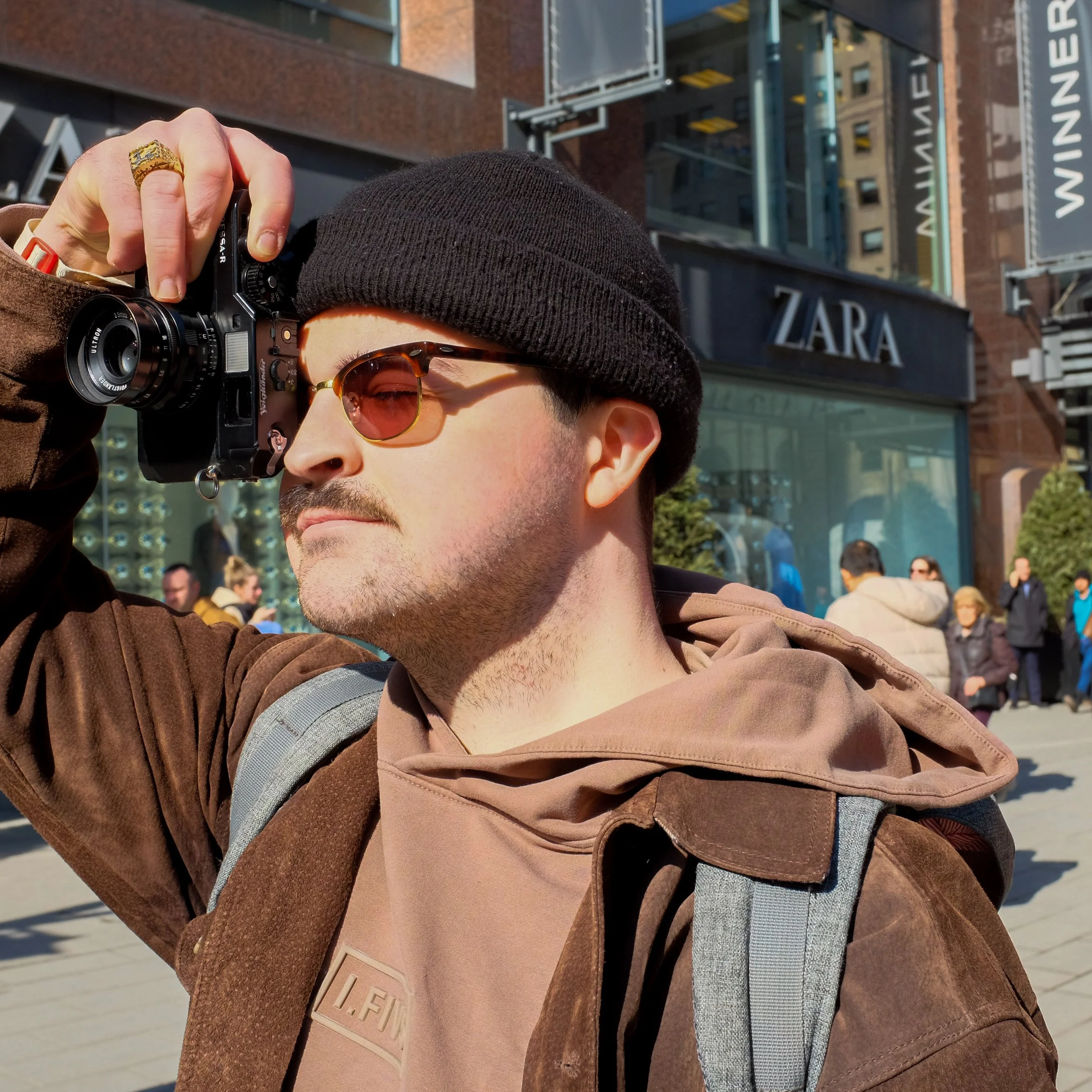 A man wearing sunglasses, a black beanie, and a brown jacket is taking a photo with a camera in an urban area in front of a Zara store.